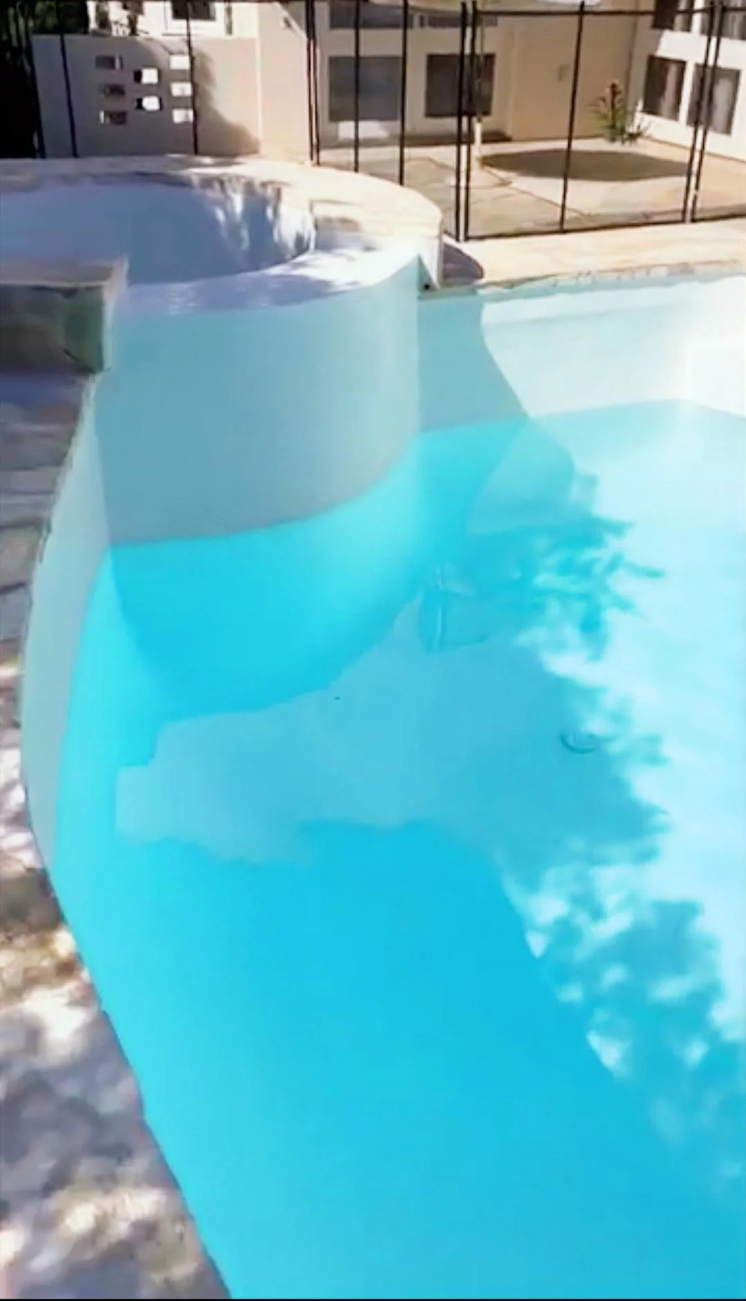 A swimming pool with a built-in hot tub, surrounded by a fence and a patio with buildings in the background.