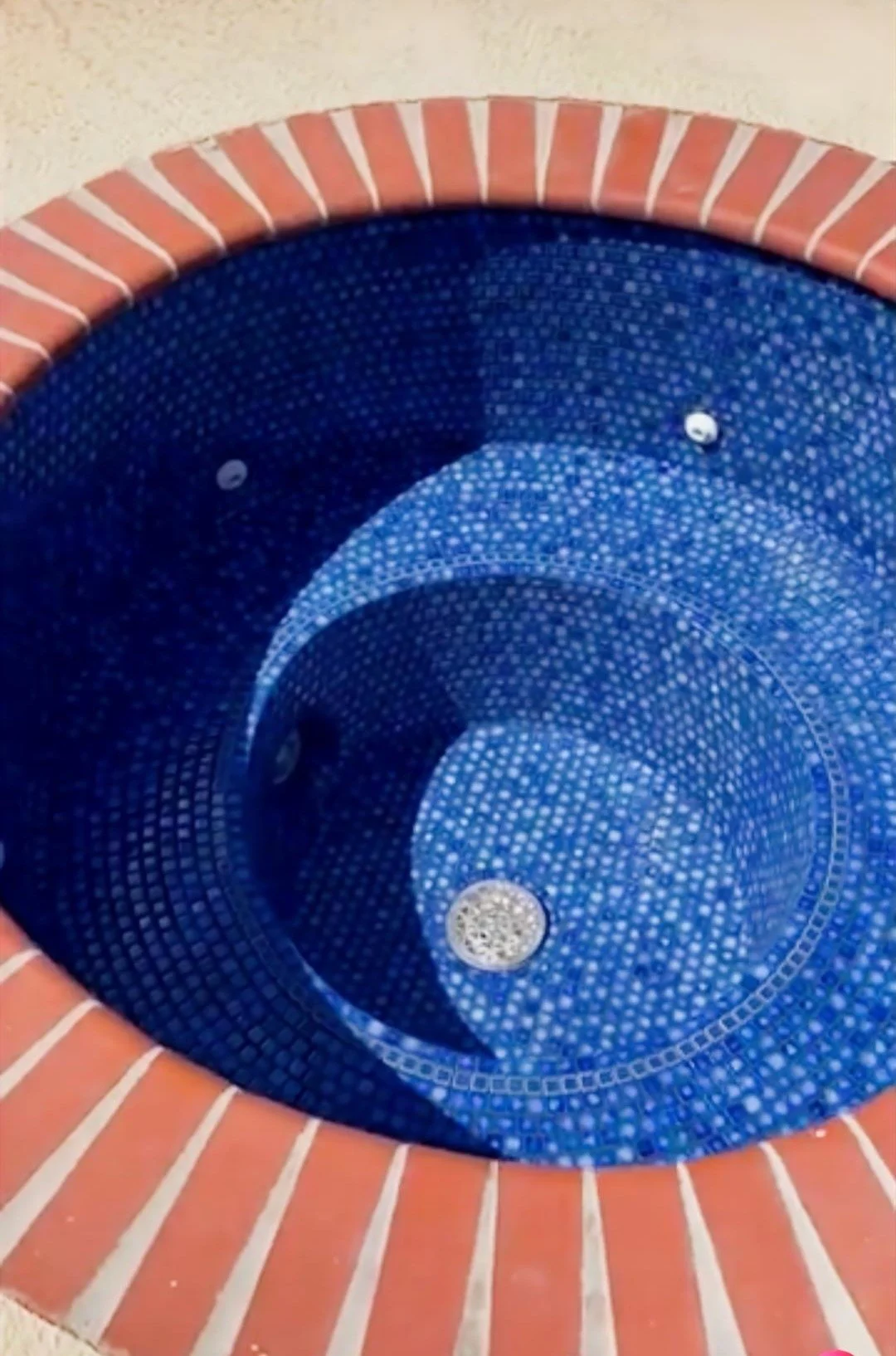 A close-up view of an empty, round, tiled swimming pool with blue mosaic tiles and a brick surround.