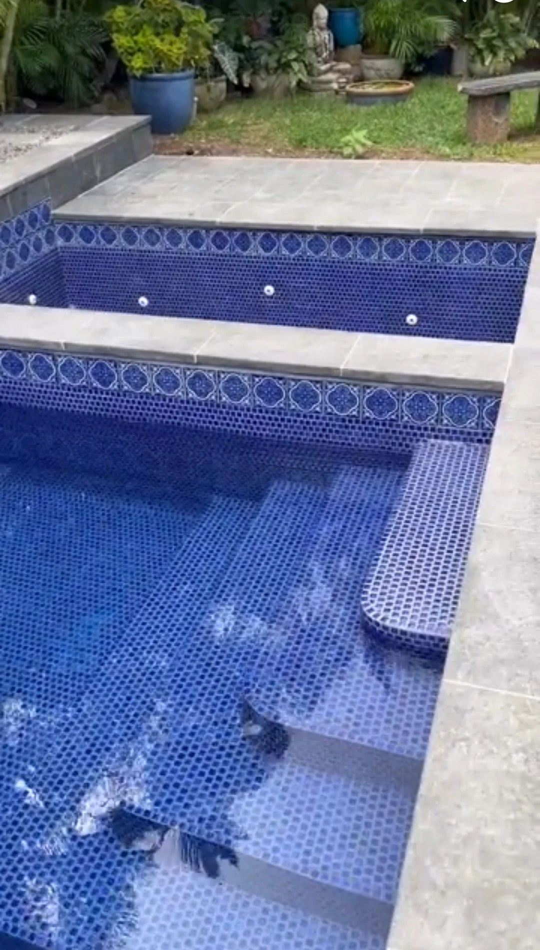 Empty swimming pool with blue mosaic tiles, surrounded by a gray stone deck, located in a backyard with green plants and stone garden decor.