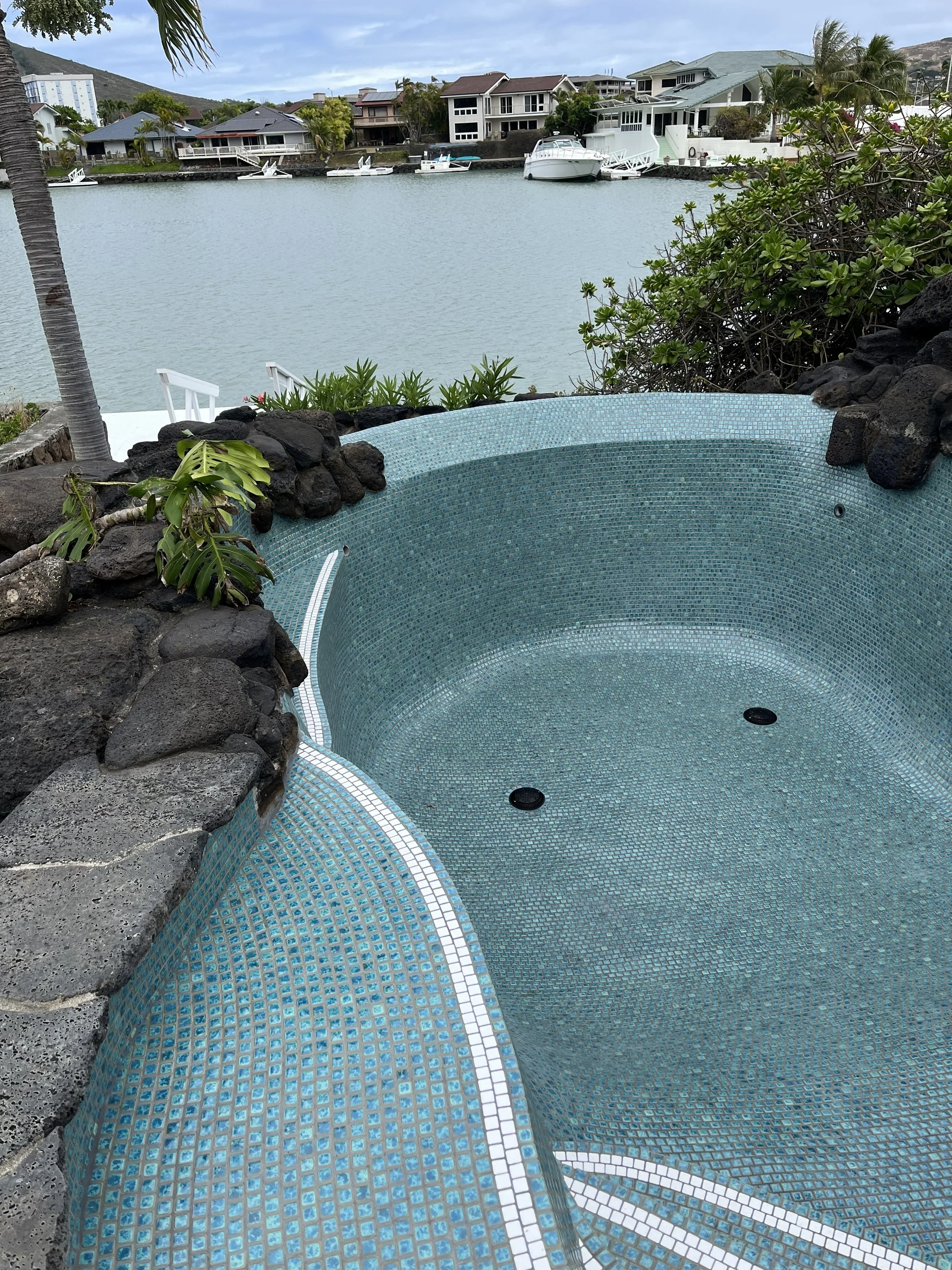 Empty mosaic hot tub overlooking a waterway with houses and boats, surrounded by greenery.
