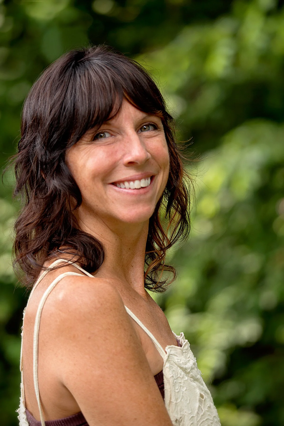 A woman with dark brown, wavy hair smiling outdoors with green foliage in the background.