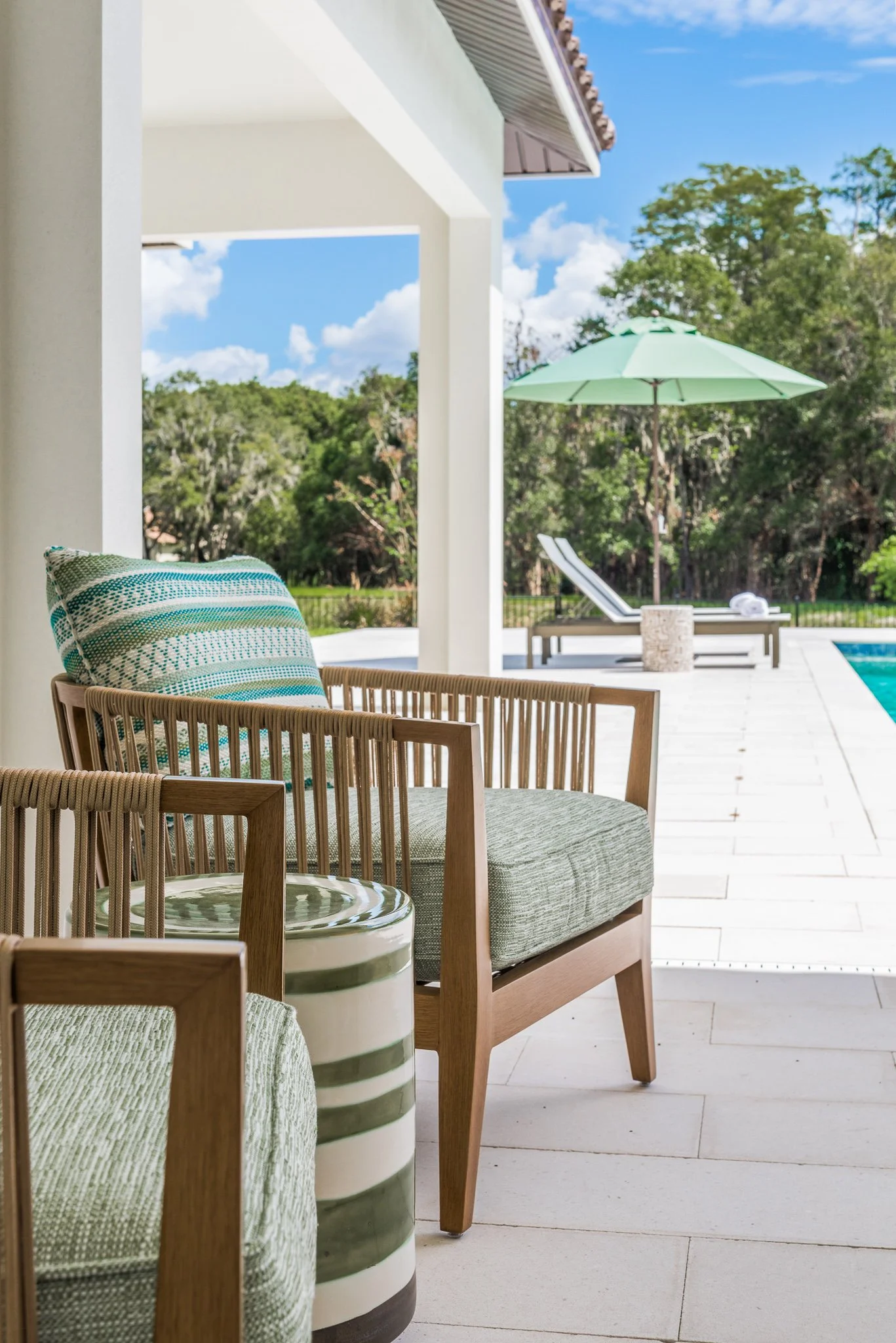 View of a backyard patio with outdoor furniture, lounge chairs, green umbrella, swimming pool, and trees in the background.