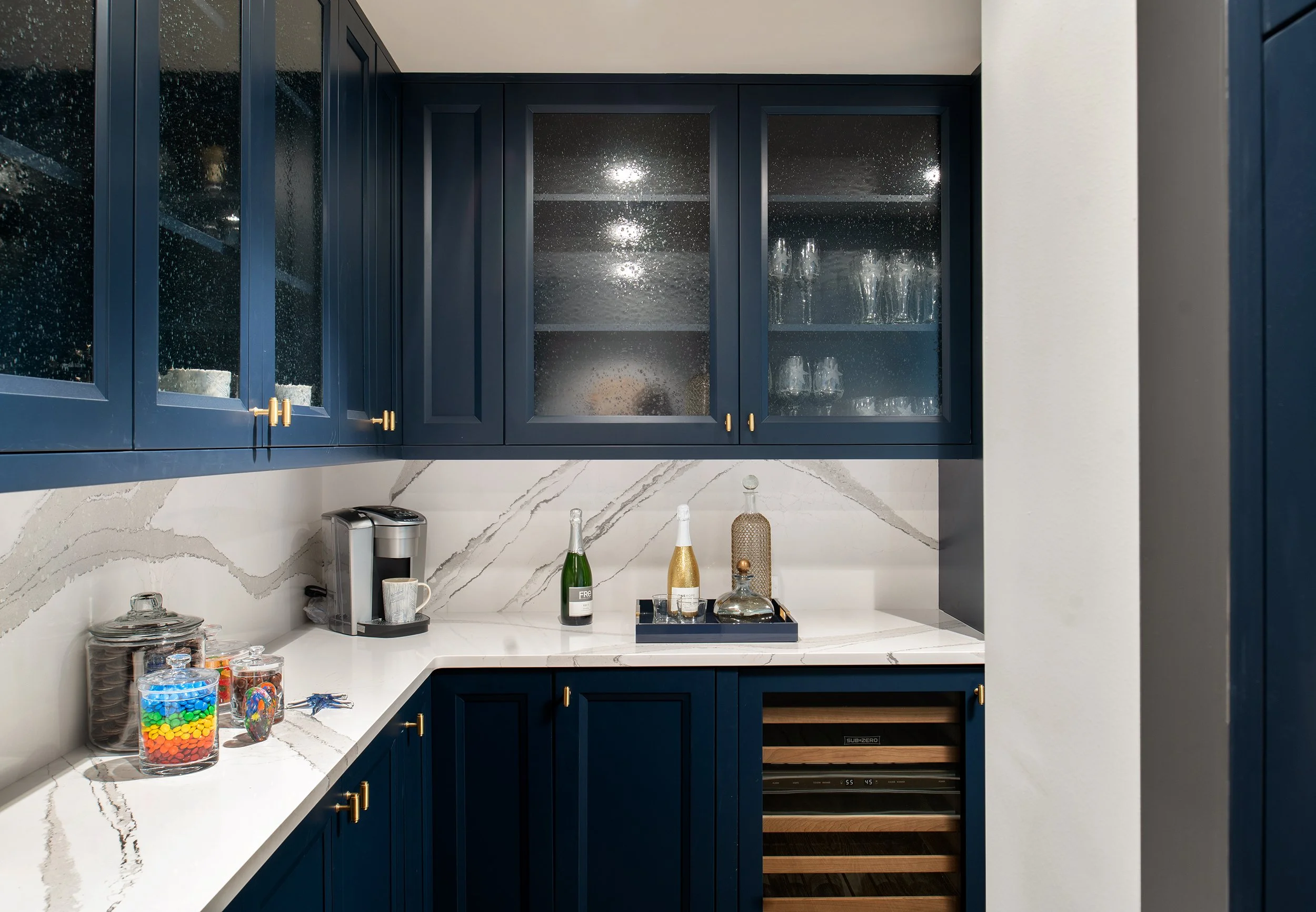 Kitchen with navy blue cabinets, white marble countertops, a coffee machine, jars with candy and cookies, and decorative bottles.