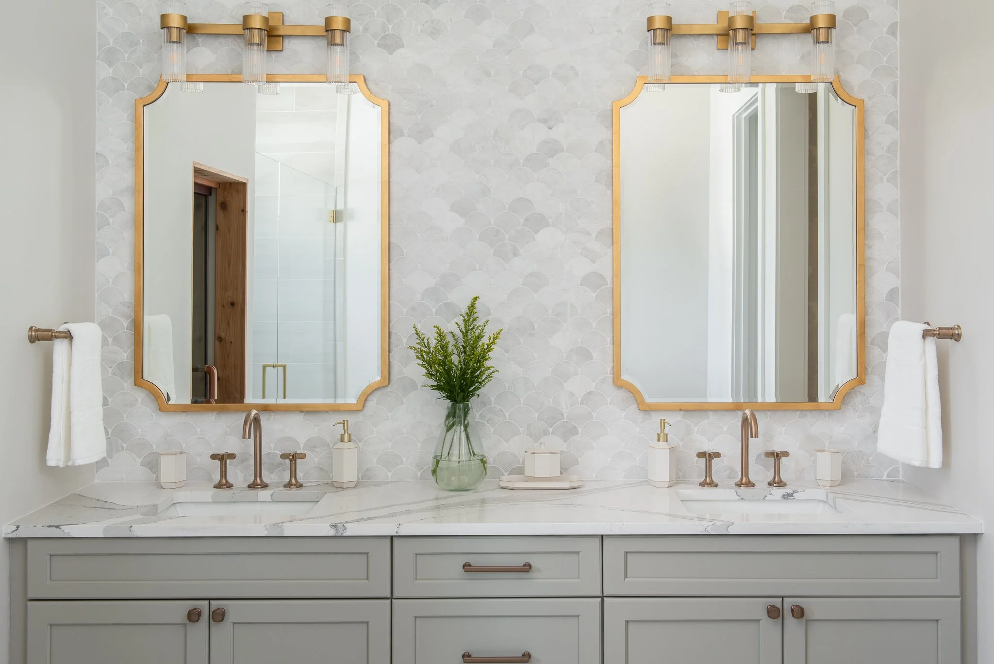 A double bathroom vanity with two large mirrors, a white marble countertop, and a vase with green foliage in the center. Each side has a faucet and soap dispenser, with white towels hanging on towel bars. The wall behind features a textured pattern w