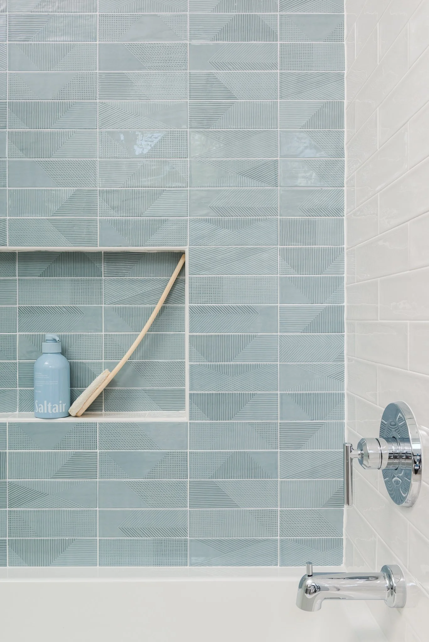 Close-up of a bathroom shower wall with blue patterned tiles, a built-in shelf holding a blue saltair bottle and a cleaning brush, and chrome shower fixtures on a white wall.
