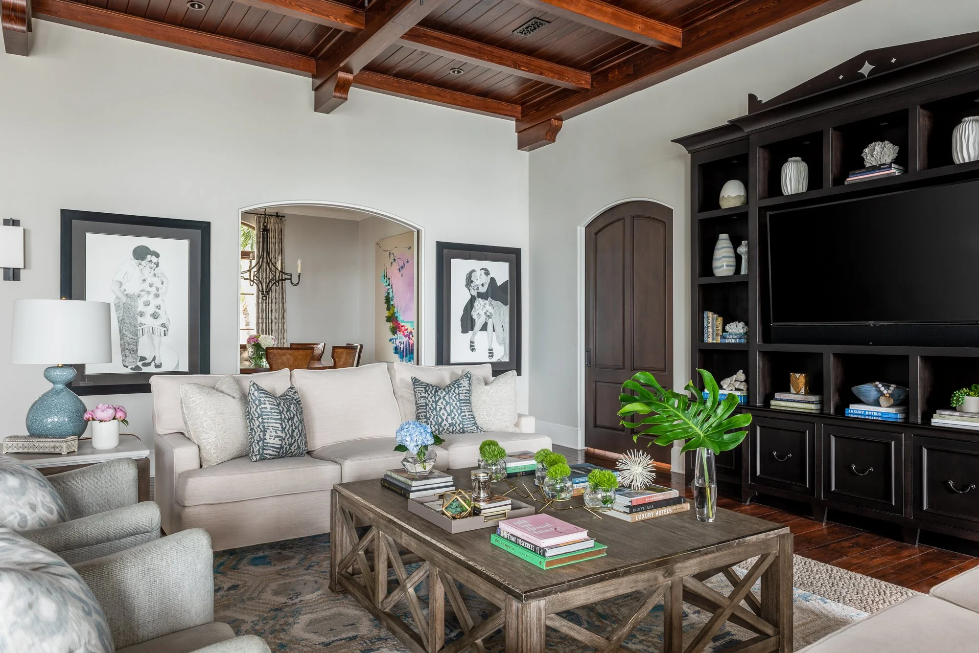 Living room with beige sofas, wooden coffee table, large black entertainment center, decorative plants, paintings, and a chandelier visible through an arched doorway.