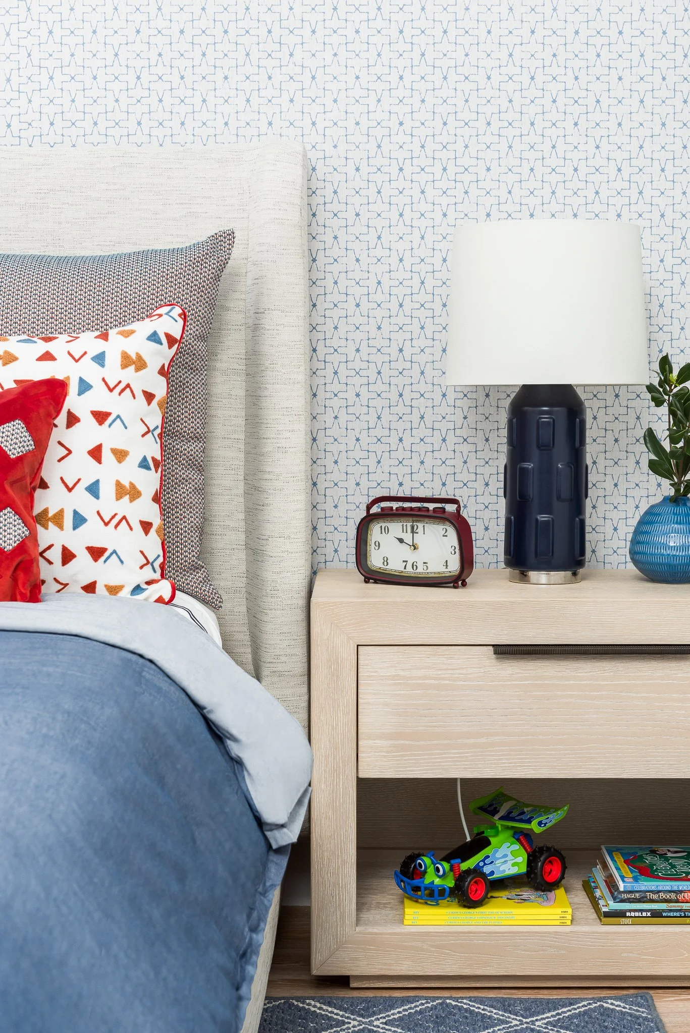 A bedroom nightstand with a white lamp, red alarm clock, and a potted plant in a blue vase. A toy car and a few books are stored on the lower shelf. Part of the bed with blue bedding and decorative pillows is visible.