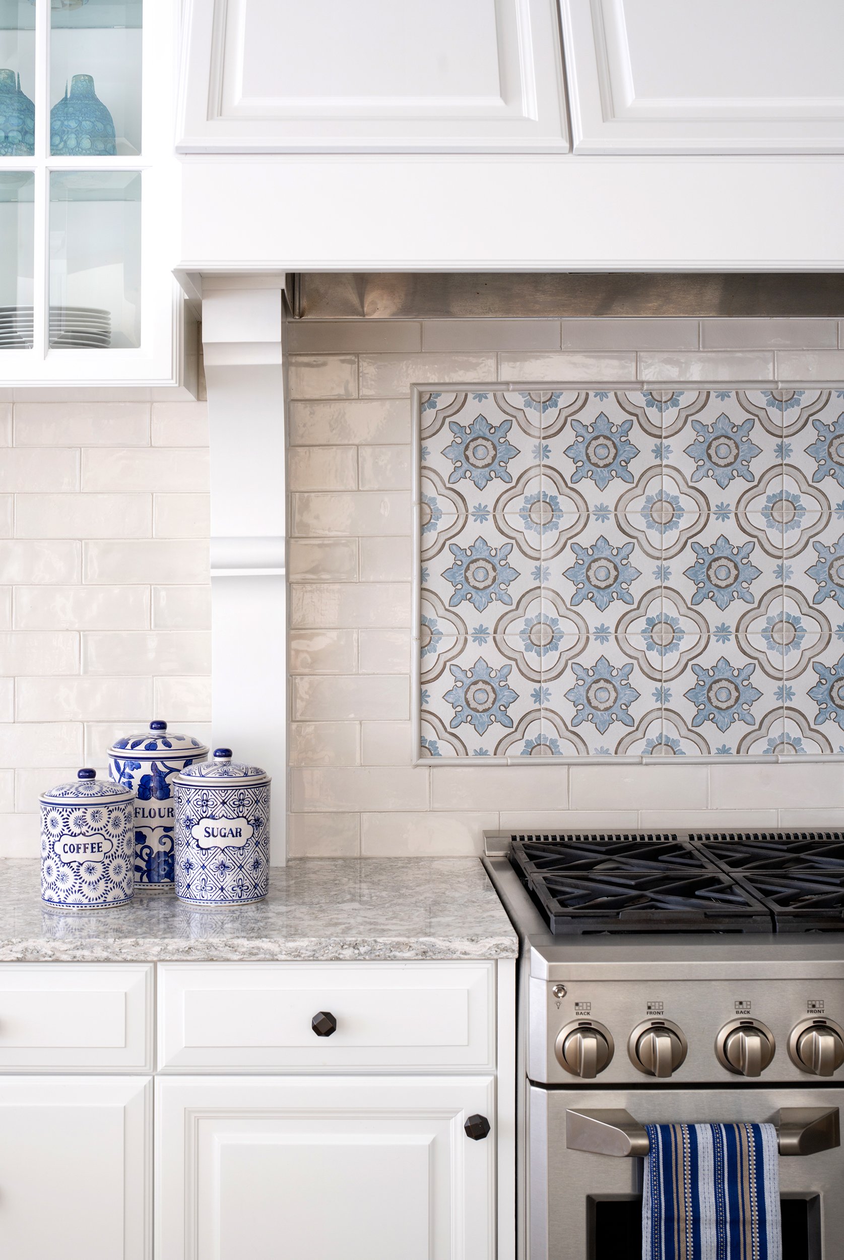 Kitchen with white cabinetry, a gray granite countertop, three blue and white ceramic containers labeled "Coffee," "Flour," and "Sugar," a stainless steel stove, and a decorative tile backsplash with a blue and gray pattern.