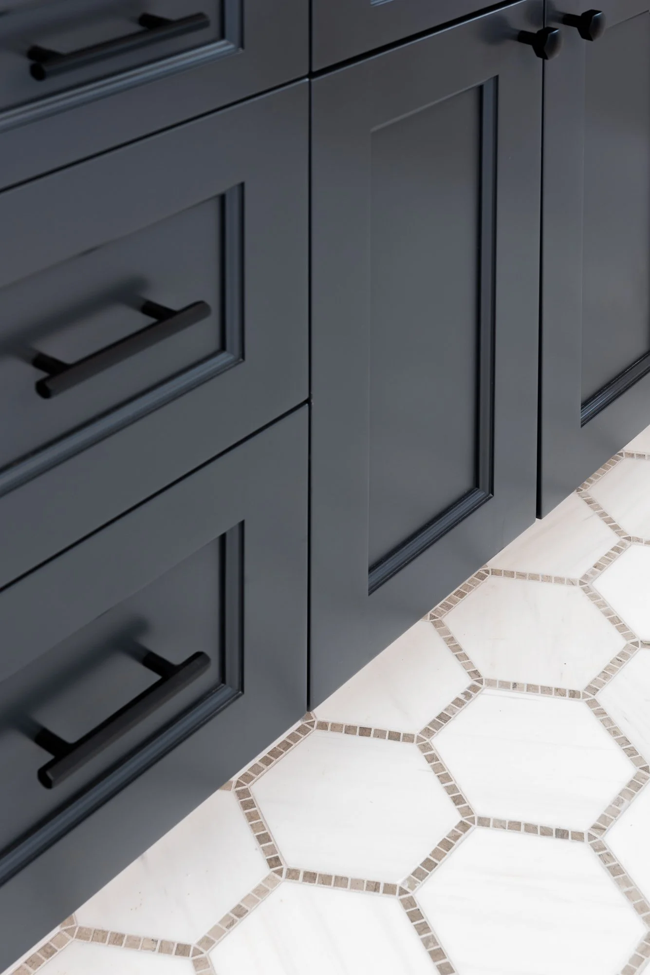 Close-up of black kitchen cabinets with black handles next to a white tiled floor with beige hexagonal border tiles.
