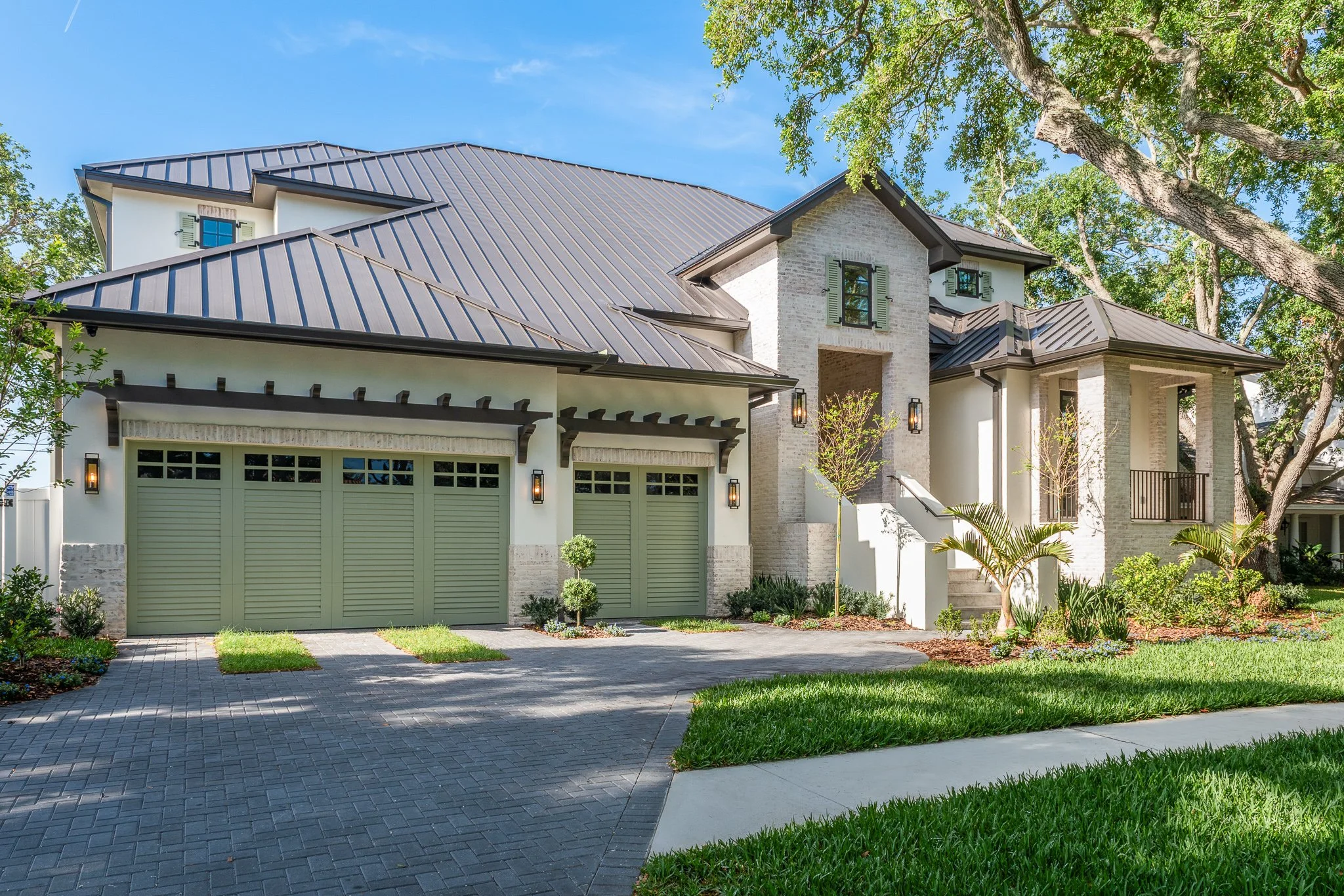 Modern two-story house with a metal roof, green garage doors, and a landscaped front yard with trees and bushes.