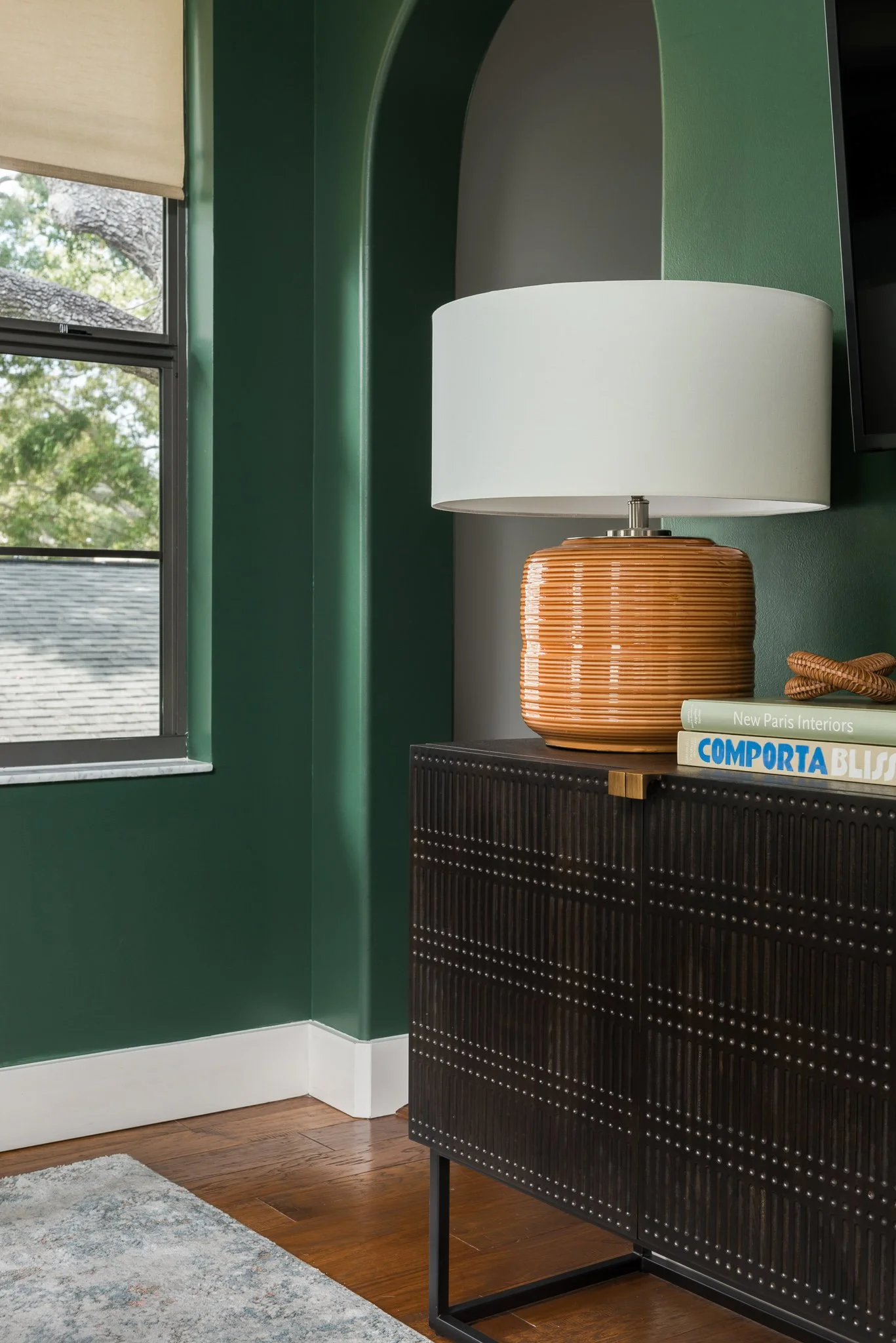 Close-up of a black wooden sideboard with textured design, topped with a large ceramic table lamp and stacked interior design books, in a room with dark green walls and hardwood floors.