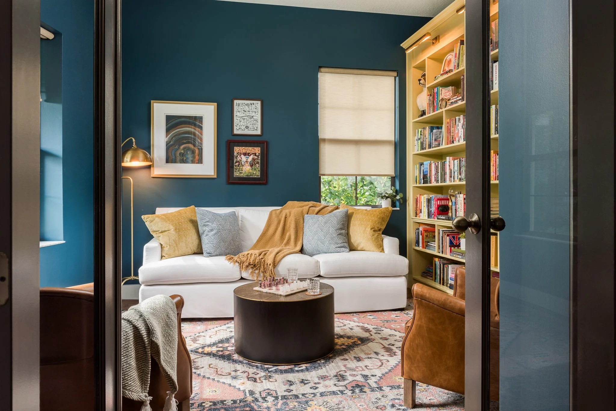 Living room with a white sofa, colorful pillows, a round black coffee table, a patterned area rug, and a yellow bookshelf filled with books, against a teal wall with art decor and a window with a beige roller shade.