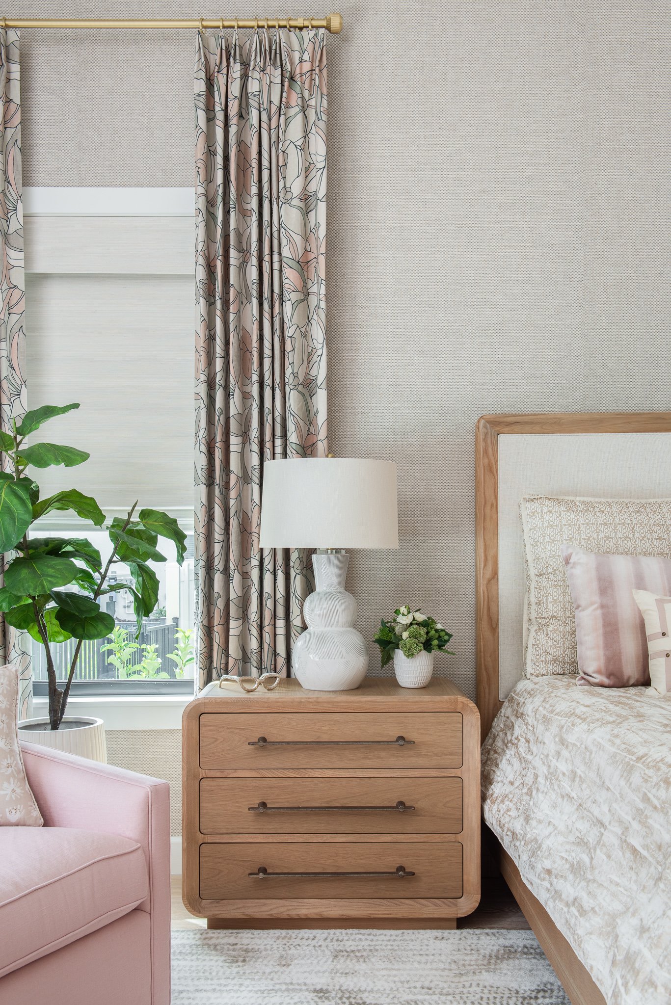 A bedroom corner featuring a wooden nightstand with a white lamp and a small potted plant, a bed with neutral bedding, a pink armchair, a green houseplant near a window with patterned curtains, and a textured wall.