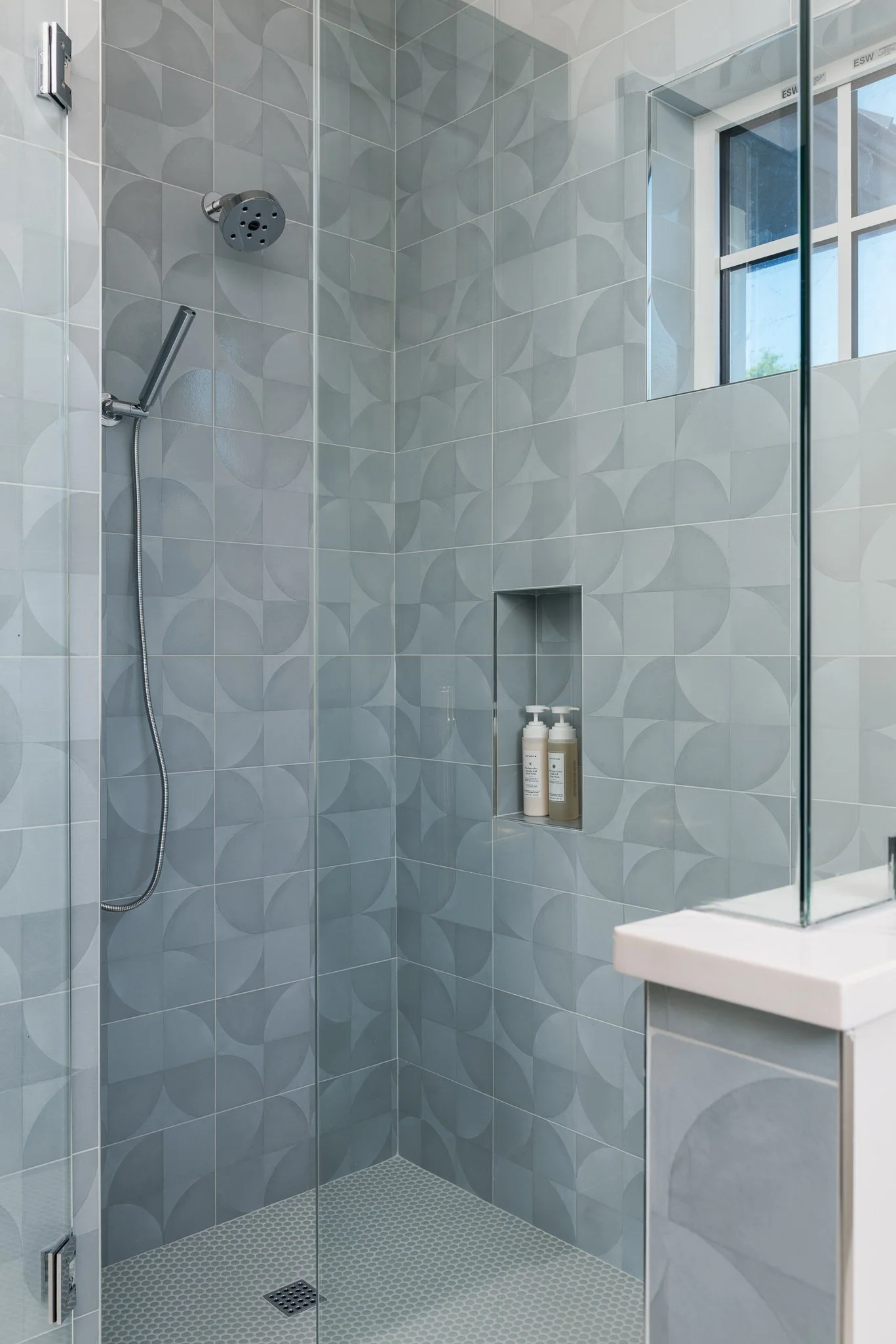 Modern bathroom shower with patterned gray tiles, a window, built-in niche with toiletries, and a glass door.