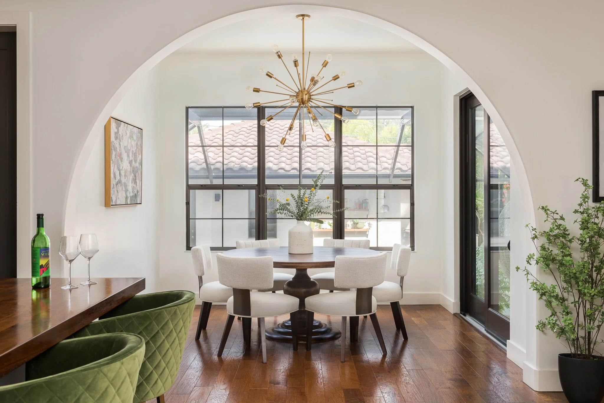 Modern dining room with a round wooden table surrounded by six white upholstered chairs, a decorative chandelier hanging from the ceiling, a large window with black framing, a white vase with greenery and yellow accents on the table, and hardwood flo