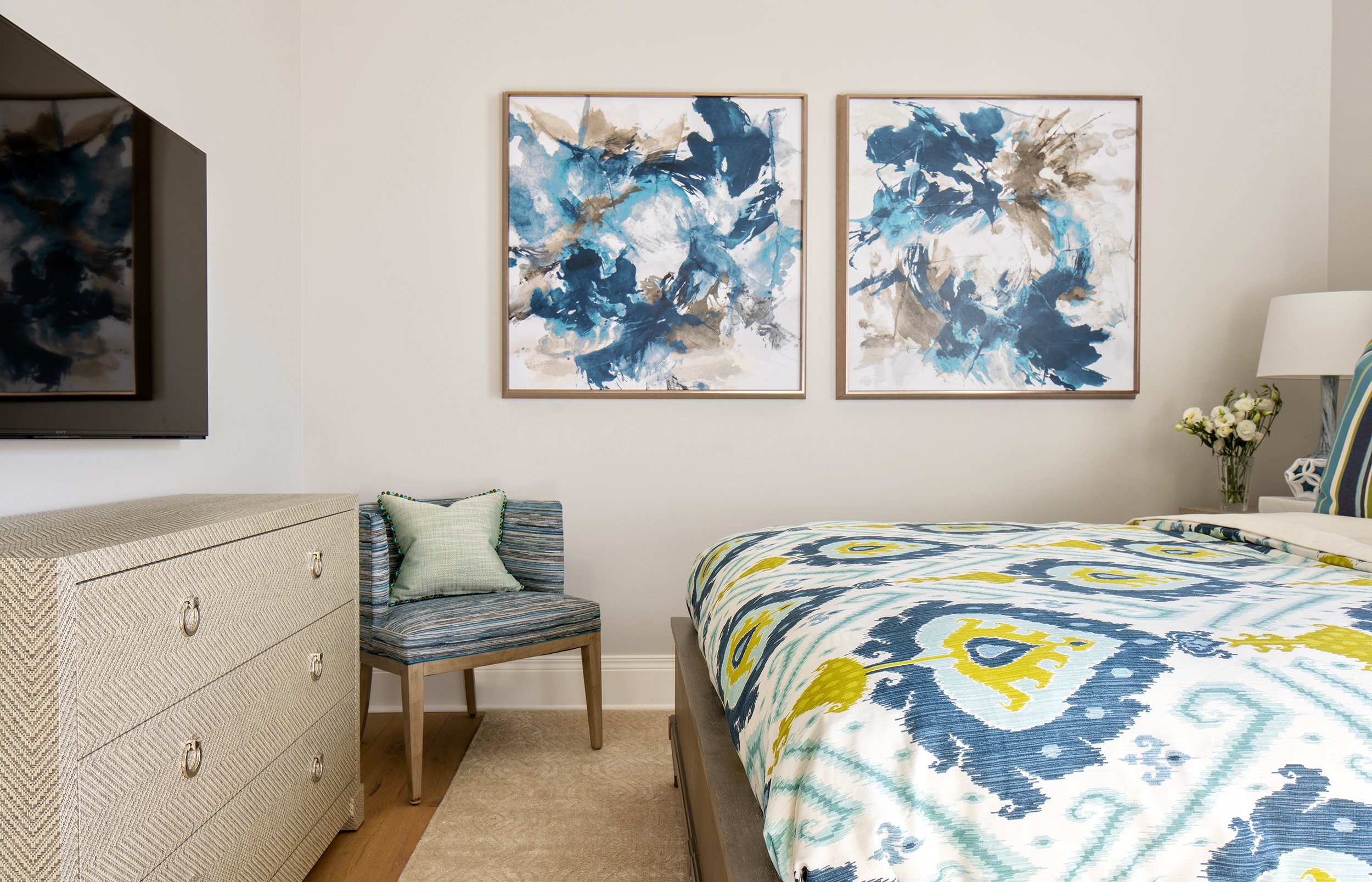 A bedroom with a bed, a side table with flowers, a chair with a pillow, and two abstract blue and beige paintings on the wall.
