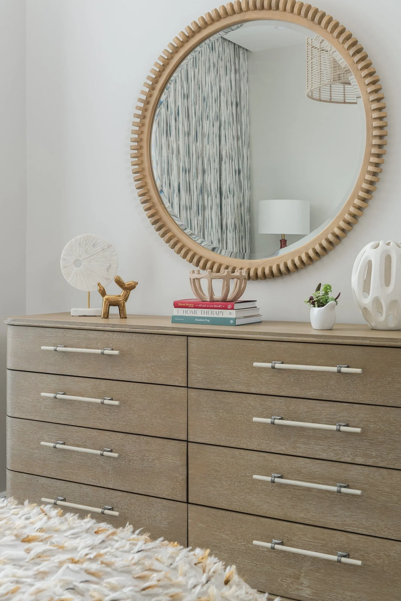 A light-colored dresser with eight drawers and modern silver handles, decorated with small decor items, a stack of books, and a small plant. A round mirror with a textured wooden frame hangs above the dresser, reflecting part of a room with a lamp an