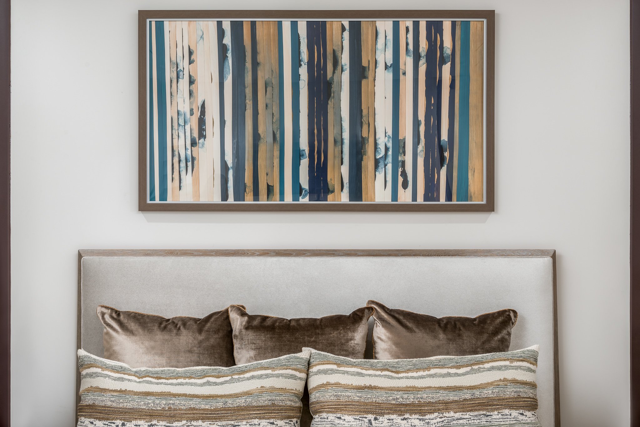 A modern bedroom with a light-colored upholstered headboard, decorative pillows, and an abstract painting with vertical blue and beige stripes hanging above the bed.