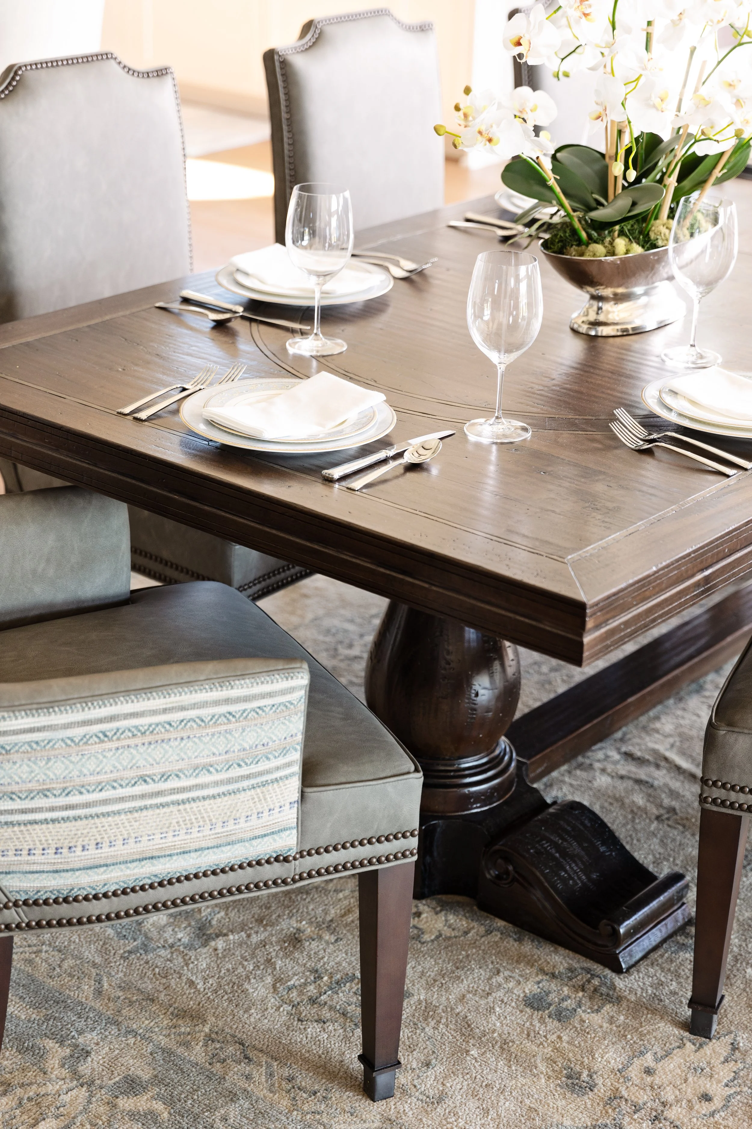Elegantly set dining table with white plates, silverware, crystal glasses, a white napkin, and a large floral centerpiece in a silver vase.