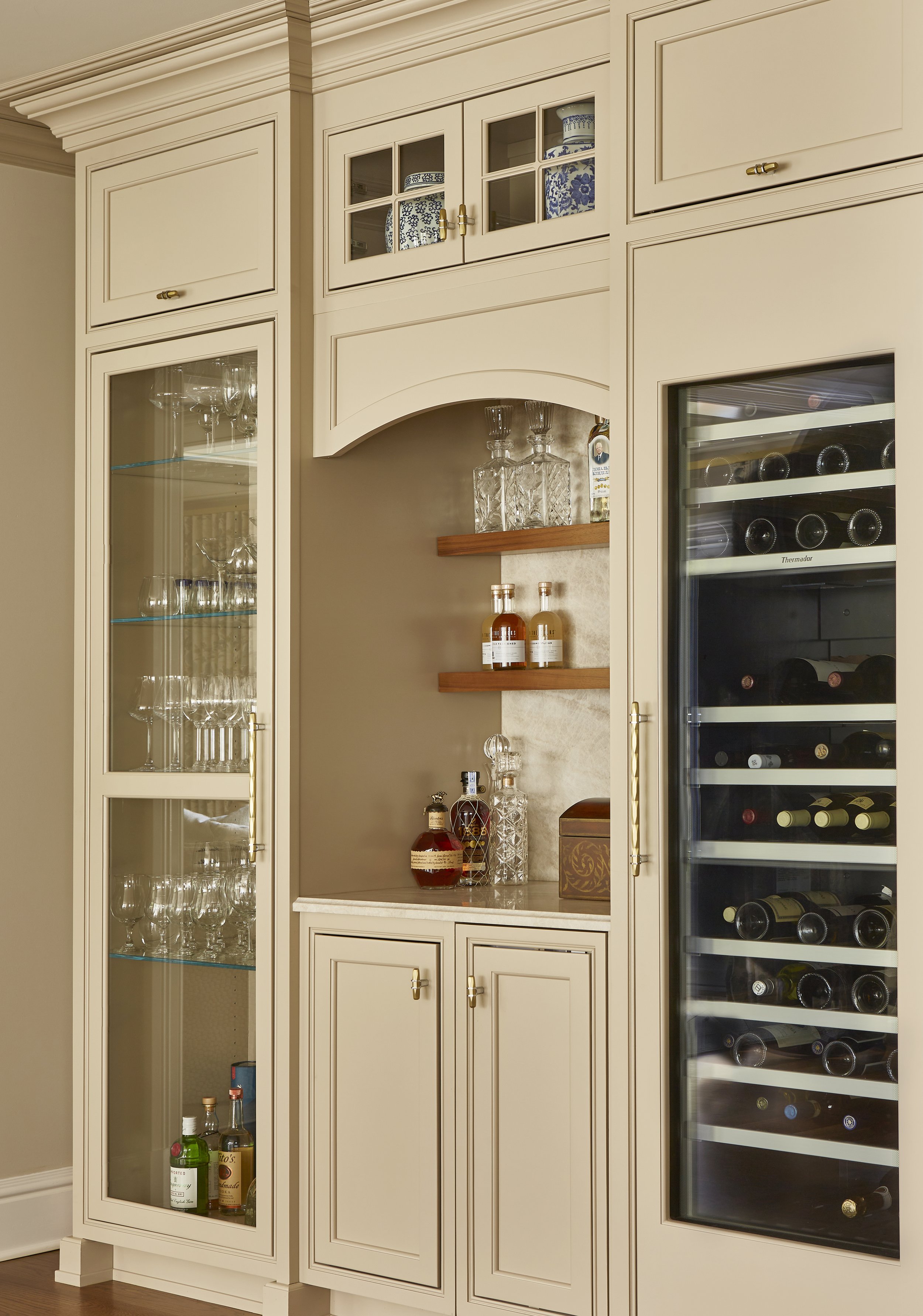 A built-in white cabinet with glass doors displaying wine glasses and liquor bottles, next to a wine refrigerator with bottles stored horizontally.