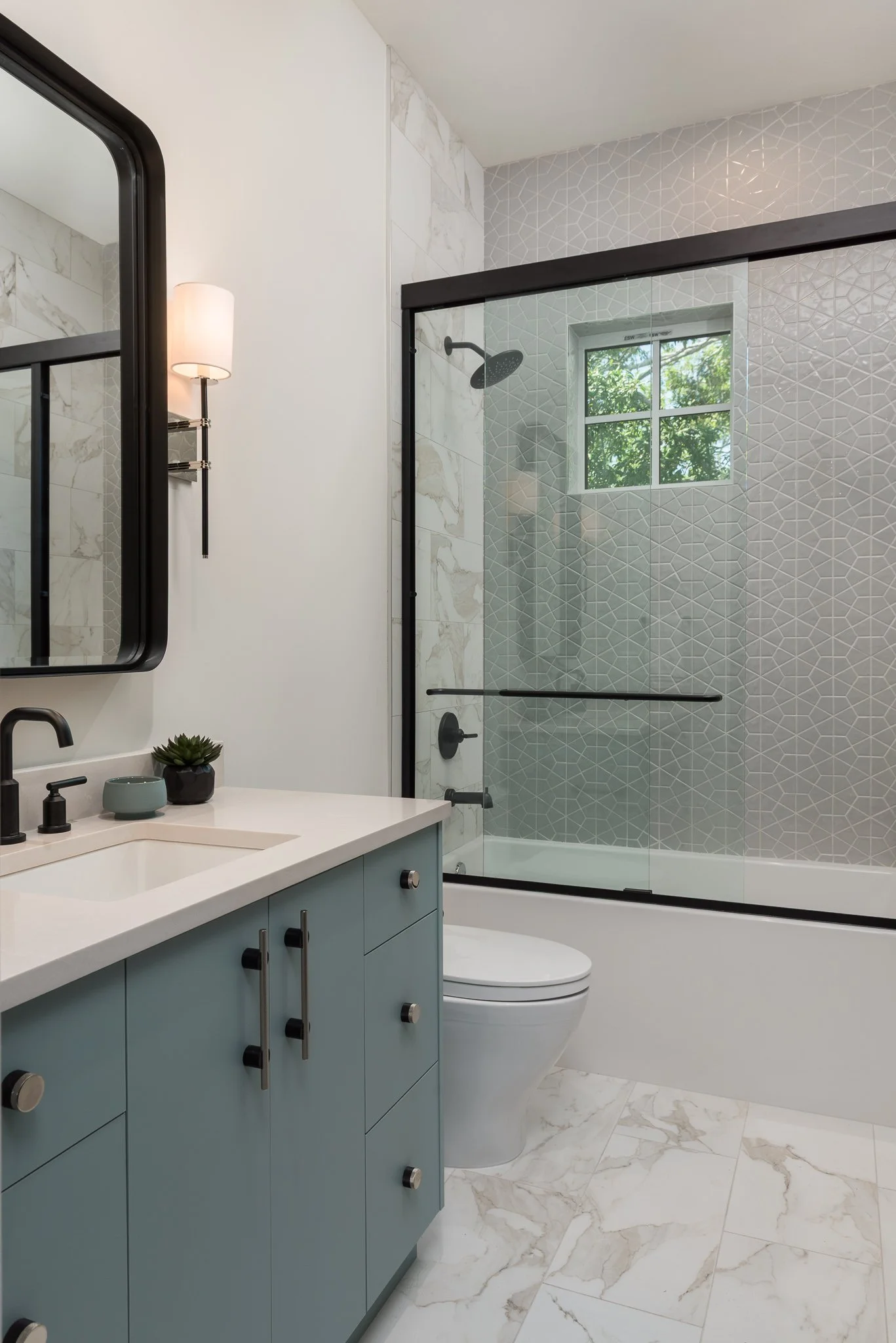 Modern bathroom with a blue vanity, a white countertop, and black fixtures. There is a mirror, a small plant, and a soap dish on the vanity. A glass shower enclosure with black trim, a showerhead, and tiled walls with geometric patterns and a window.