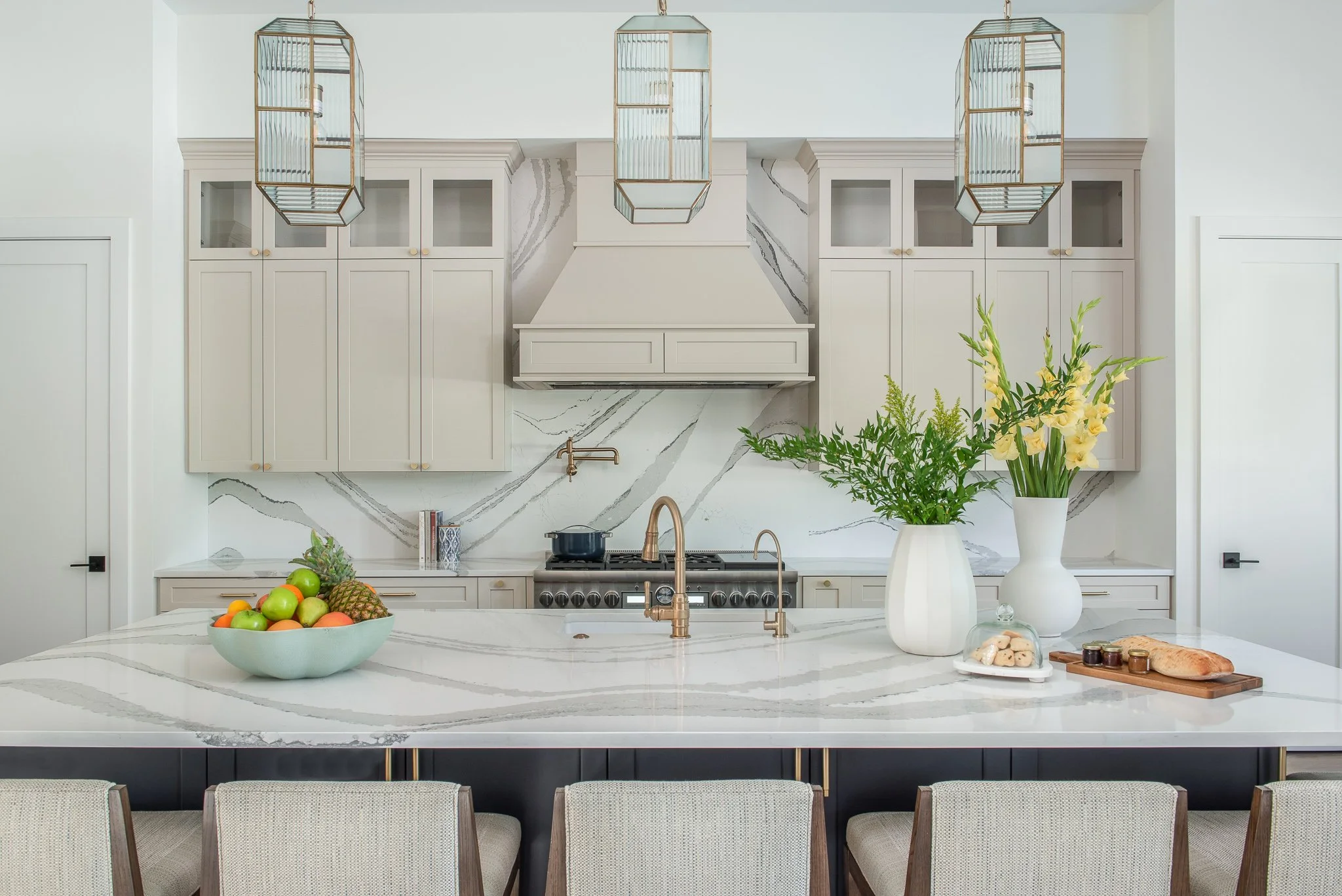 Modern kitchen with white marble island, light gray cabinets, a stove, large plants, a bowl of fruit, and hanging light fixtures.