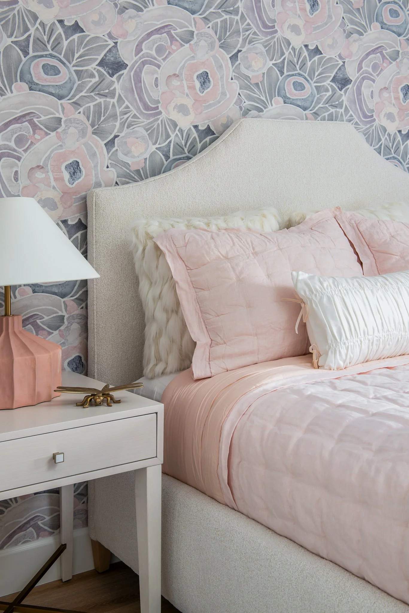 A bedroom corner featuring a light beige upholstered bed with pastel pink bedding and pillows, including a textured white bolster pillow. A white nightstand with a square drawer and brass pull holds a pink ceramic lamp with a white shade, a brass dec