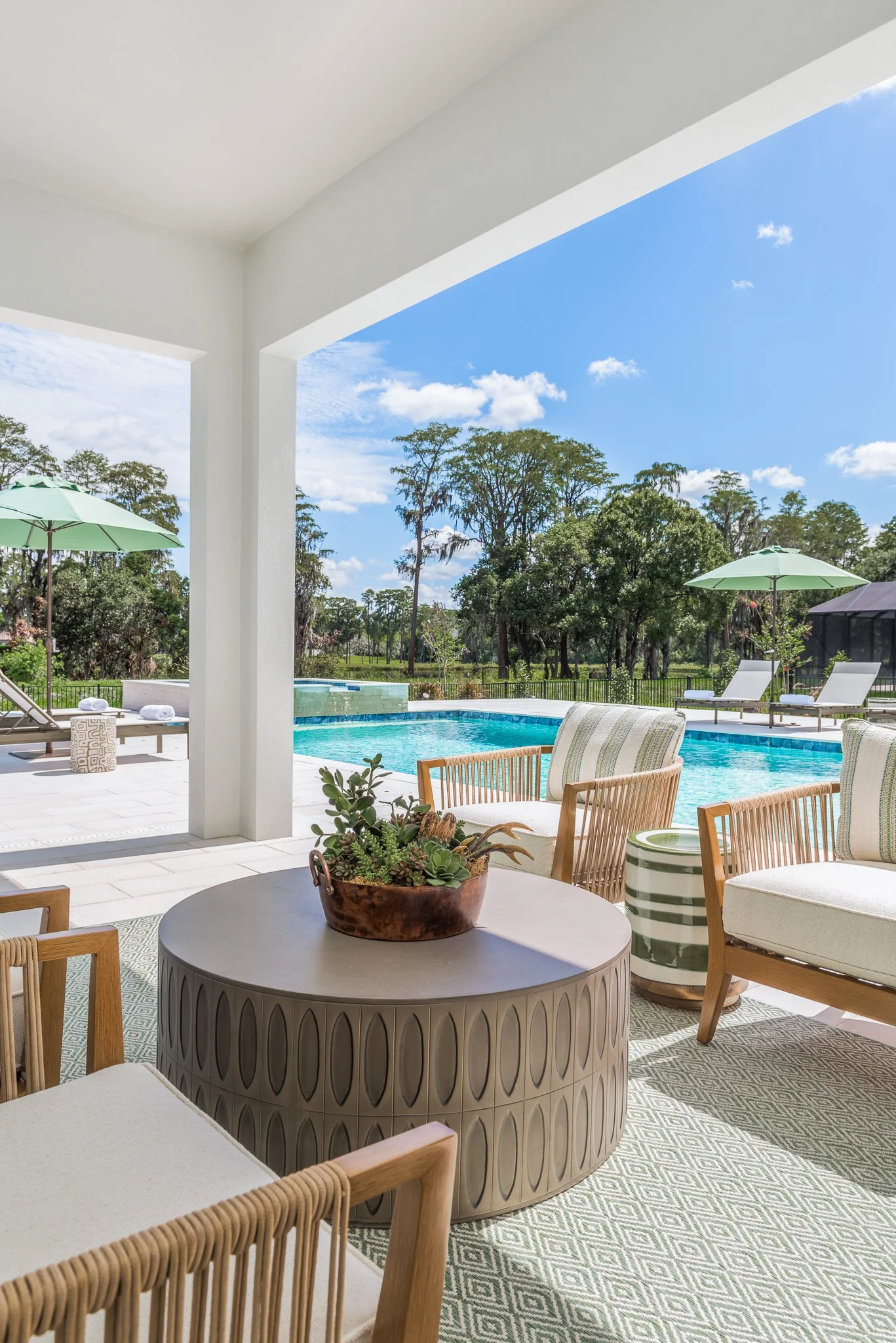 View of an outdoor pool area with lounge chairs and umbrellas, surrounded by trees and a partly cloudy sky.