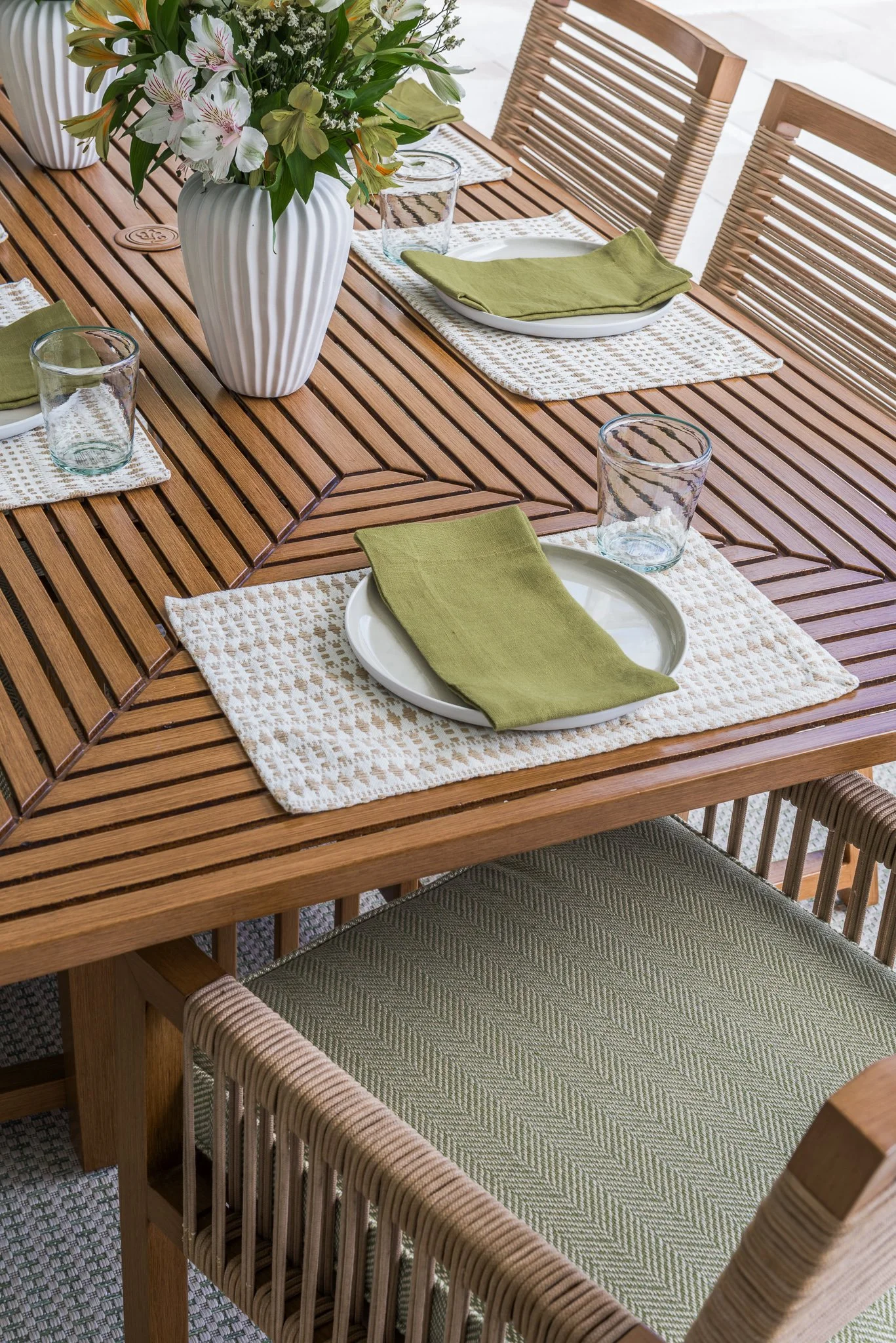 A wooden dining table set for four with white plates, green napkins, and clear glasses, decorated with a large white vase of flowers, with wicker and wooden chairs.