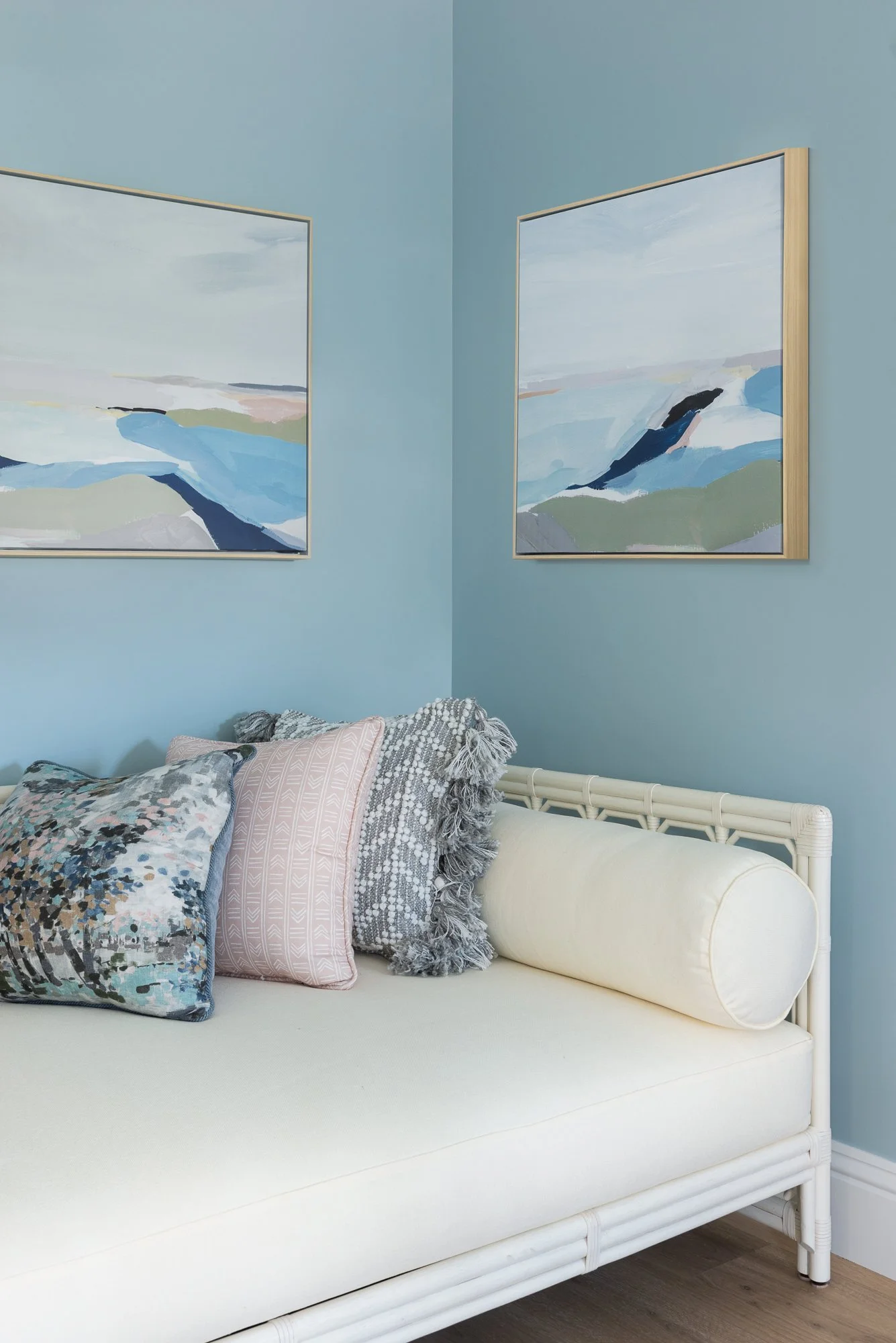 Living room corner with light blue walls, abstract landscape paintings, and a white daybed with decorative pillows.