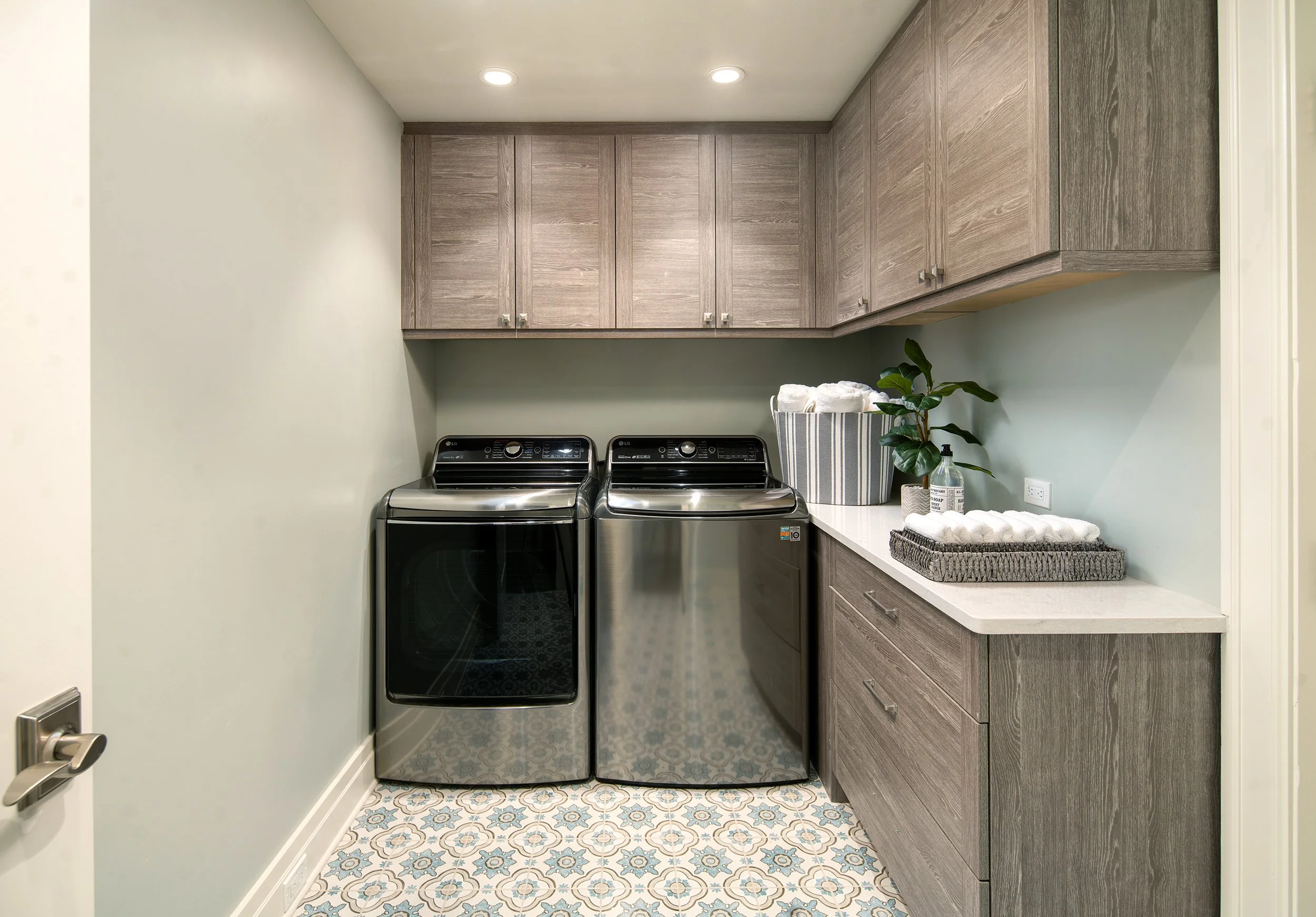A laundry room with a washing machine and dryer, wooden cabinets, a countertop with a plant, towel, soap dispenser, and decorative skirt on baskets.