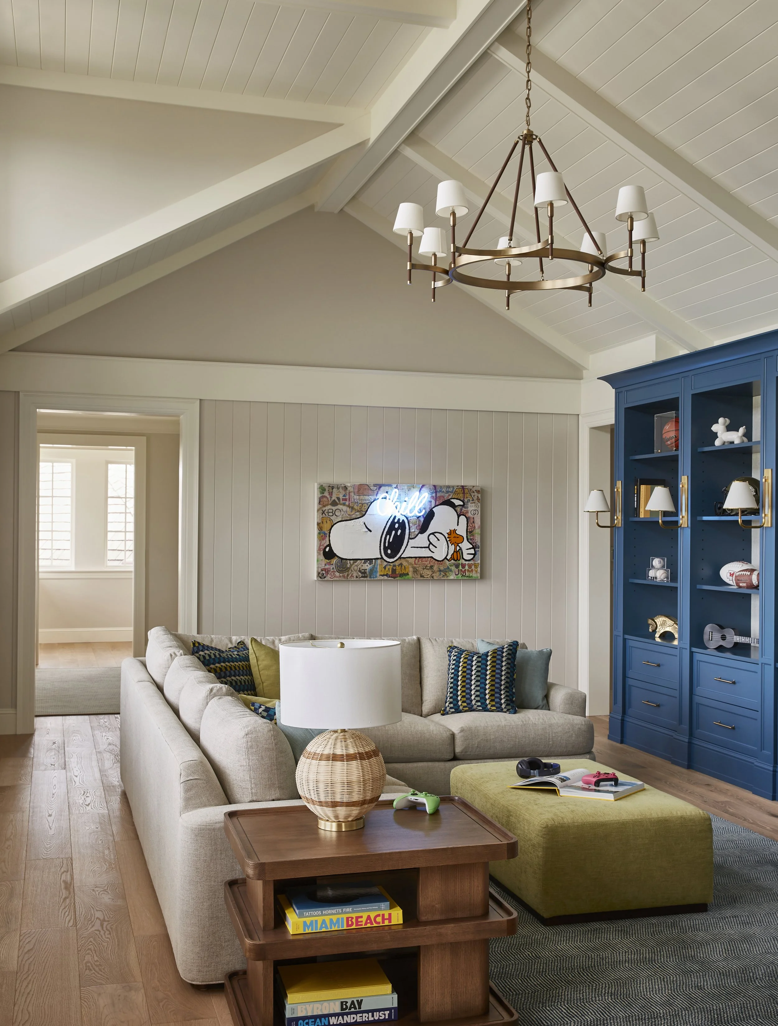 Living room with high vaulted ceiling, beige sectional sofa with colorful pillows, blue built-in bookshelf, modern art featuring Snoopy, and a wooden coffee table with books and gaming controllers.