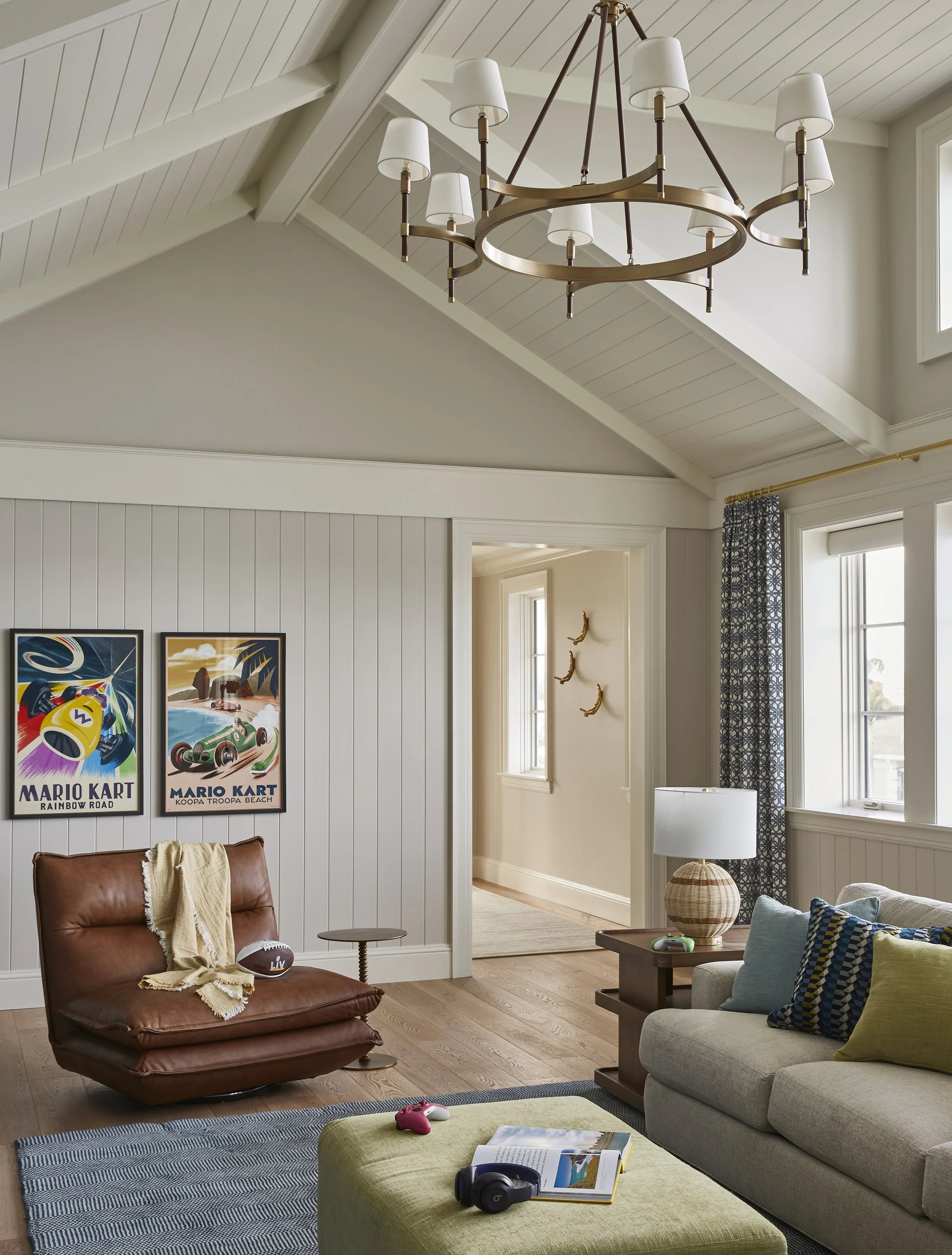 Living room with a high vaulted ceiling and white paneling, modern chandelier, armchair with a blanket, wall art posters, window with patterned curtains, sofa with pillows, side table with lamp and decorative ball, hardwood flooring, and a rug.