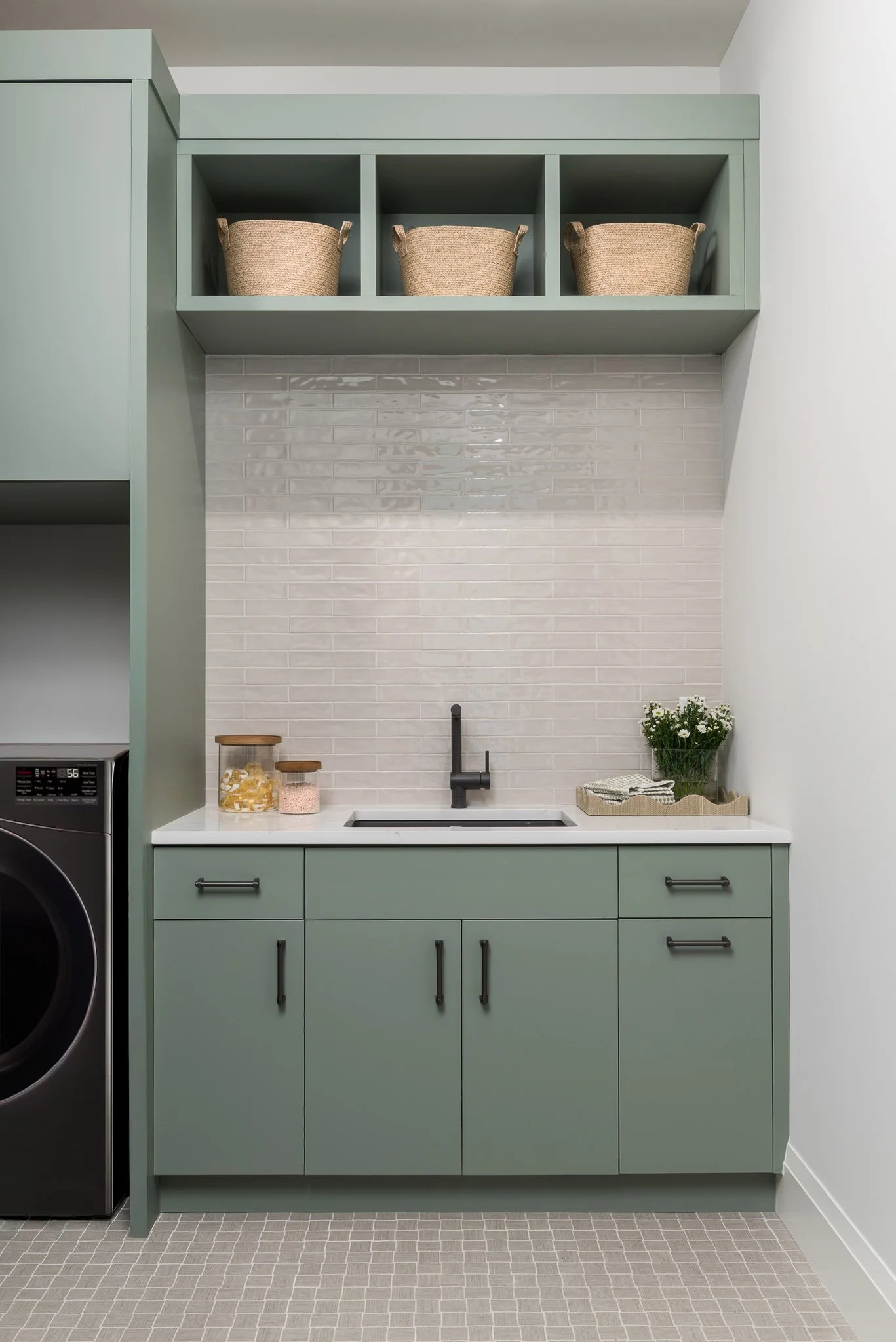 Modern laundry room with green cabinetry, a small sink, black faucet, decorative jars, and a basket with white flowers on the countertop. An open shelf with baskets above the sink.