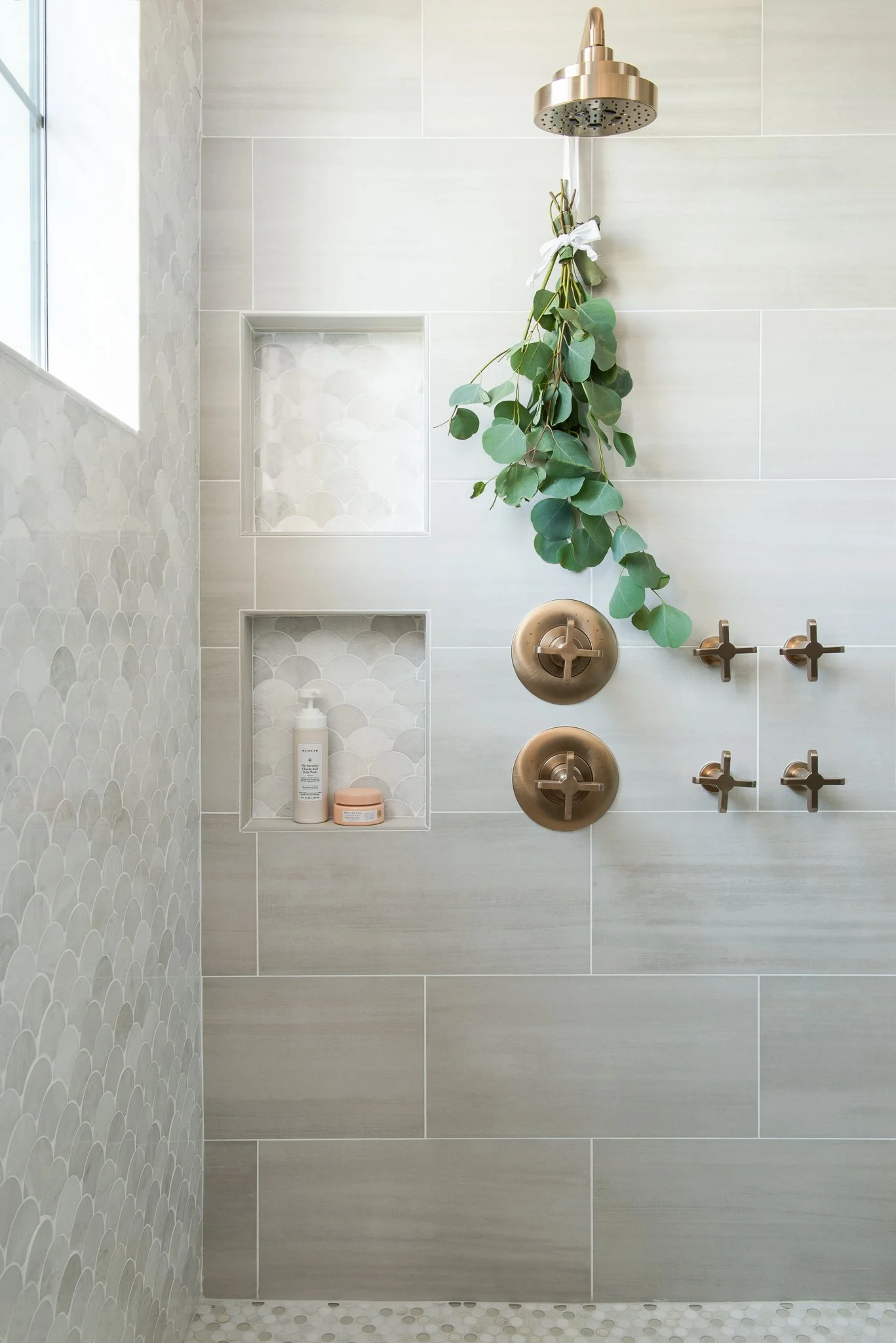 Modern shower with beige tile walls, built-in niche with soap and lotion, hanging eucalyptus, and gold-colored fixtures.