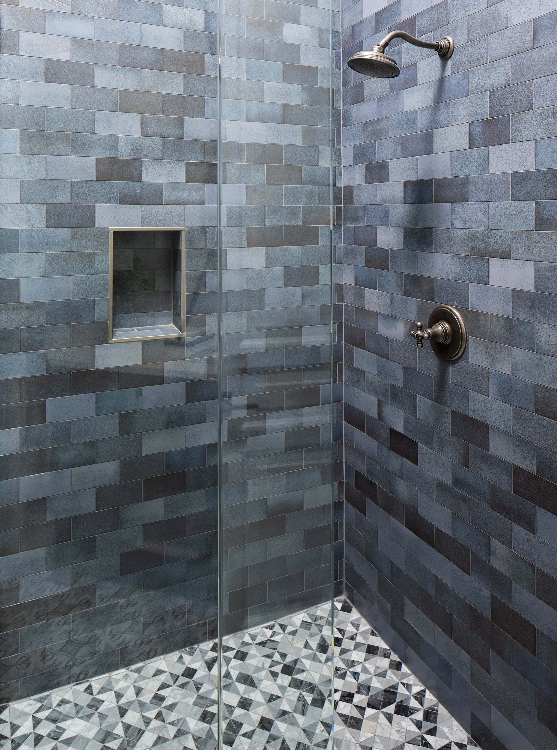 Image of a modern walk-in shower with dark gray tiled walls, a built-in niche for toiletries, a round showerhead, and a handheld control, with a patterned black and white tiled floor.