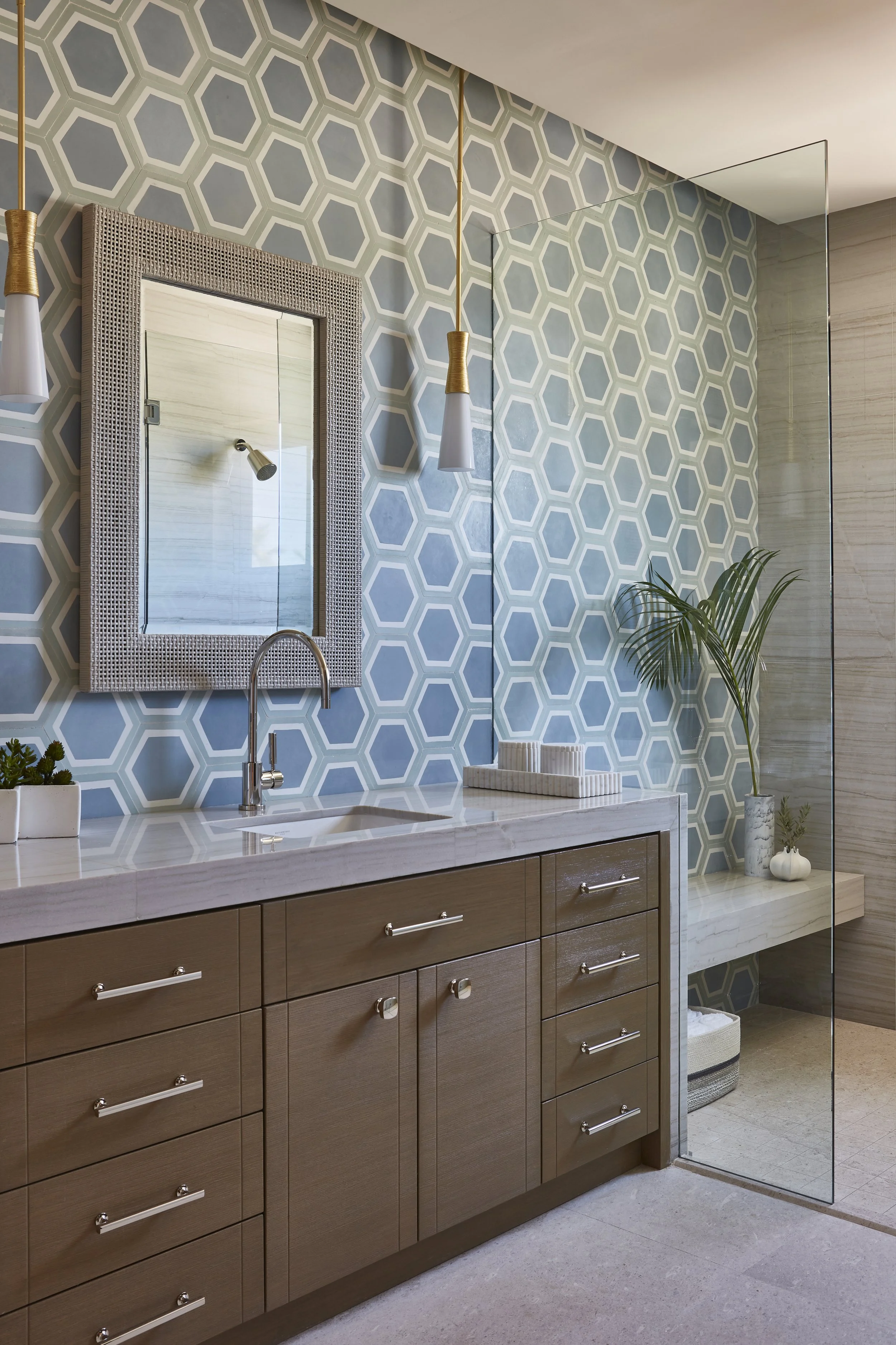 Modern bathroom with hexagonal patterned wallpaper, wooden cabinetry, marble countertop, a mirror with a textured frame, pendant lights, and a decorative plant.