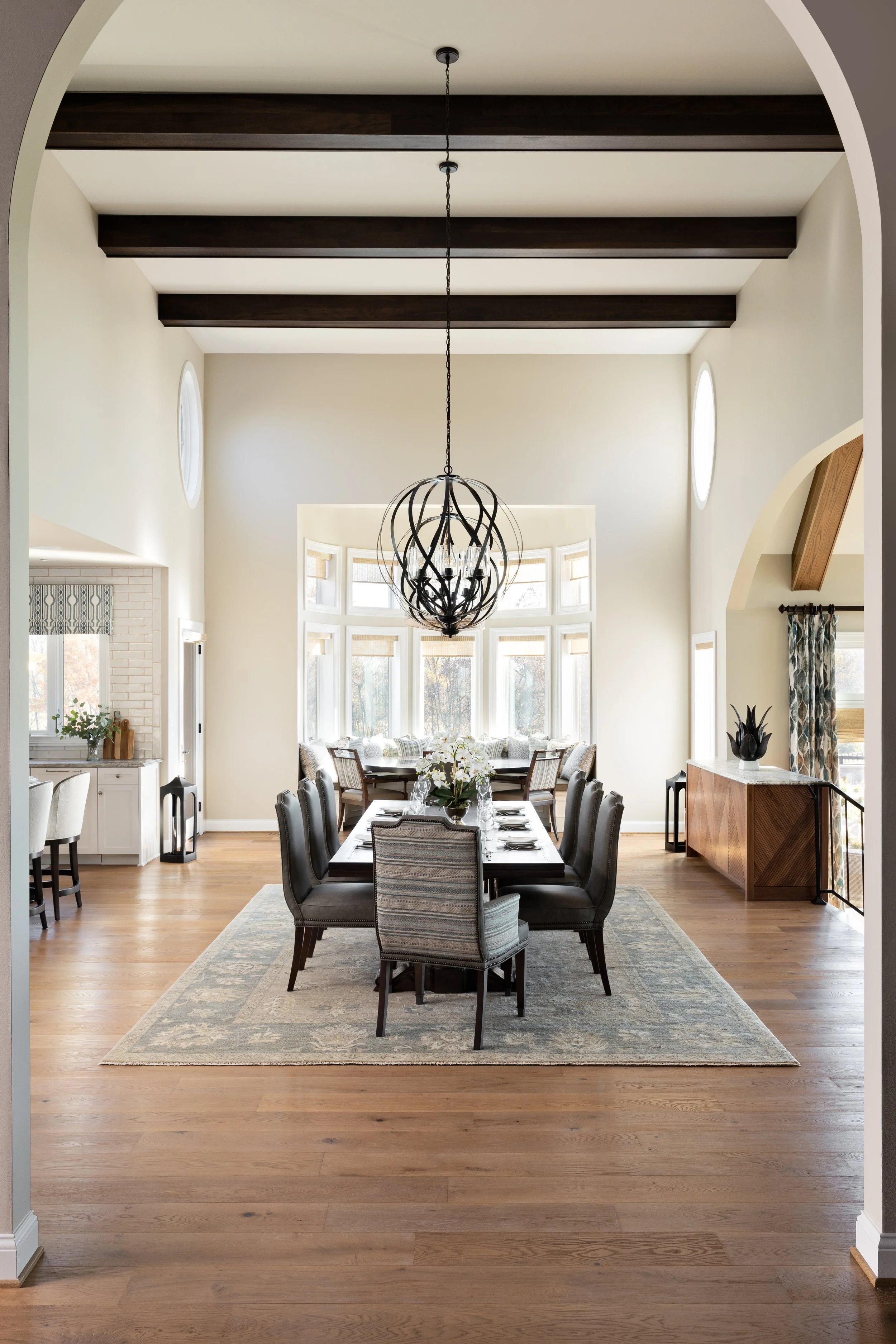 Dining room with a long wooden table, eight chairs, a chandelier, hardwood floors, and large front windows with a bay window seat.