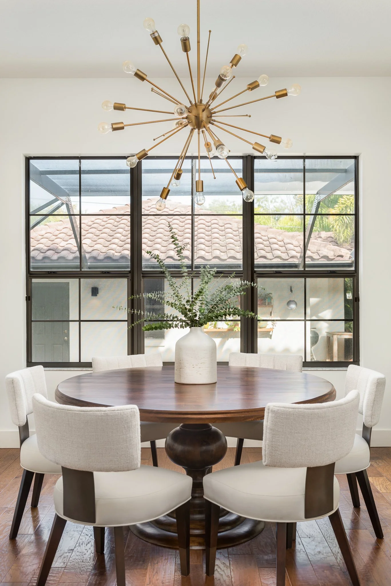 A round wooden dining table with a large ceramic vase holding greenery in the center, surrounded by six white upholstered chairs in a bright room with large windows and a modern chandelier overhead.