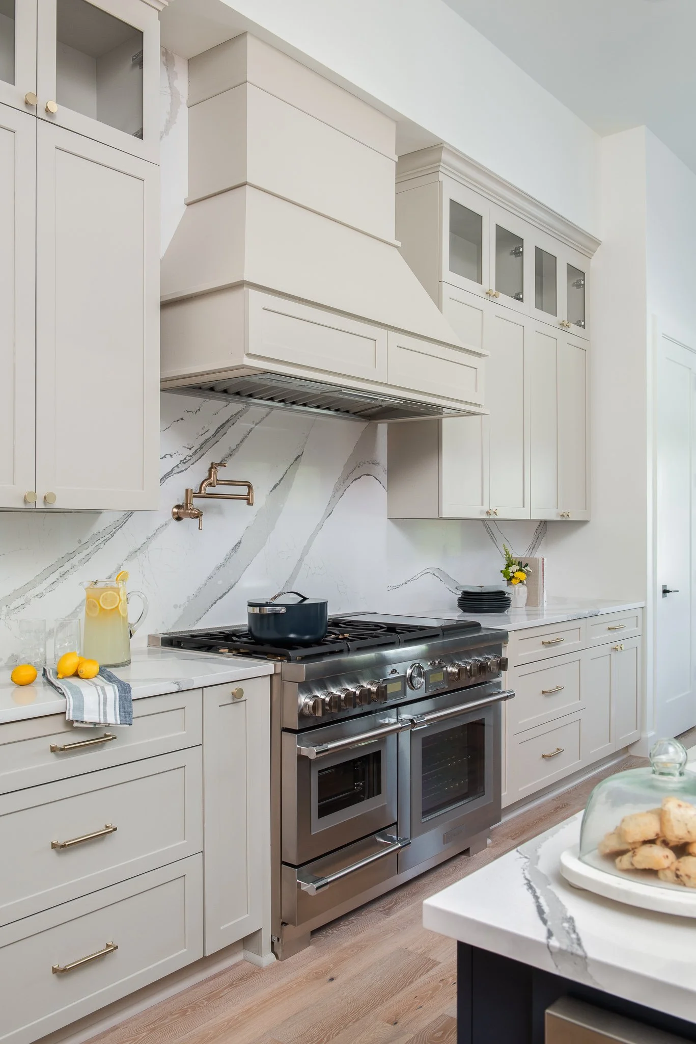 Modern kitchen with white cabinets, marble backsplash, stainless steel oven, black pot on stove, pitcher of lemon water, and a plate of cookies on a marble island.