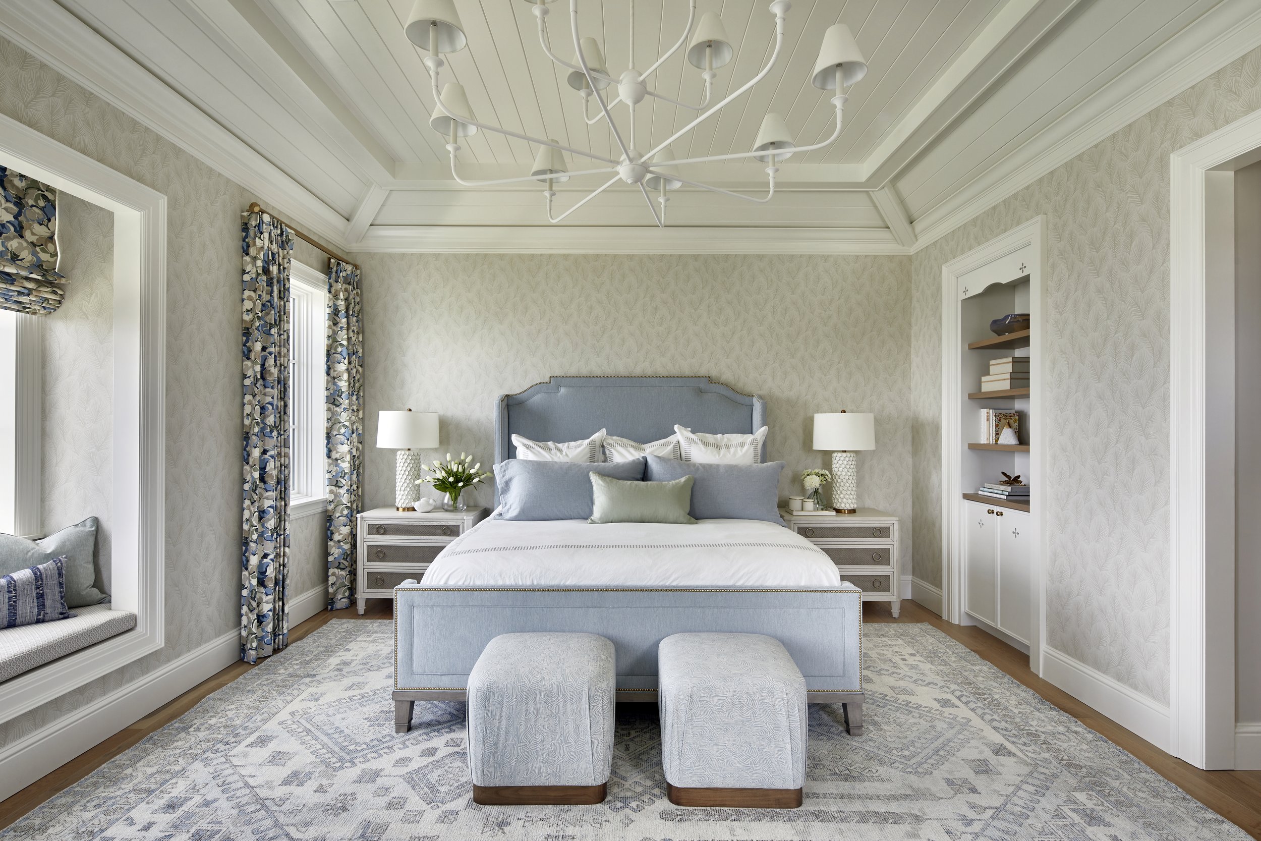 Elegant bedroom with a queen-sized bed, light blue upholstered headboard, multiple decorative pillows, two matching nightstands with lamps, a built-in bookshelf, window seat, and a white chandelier against patterned wallpaper.