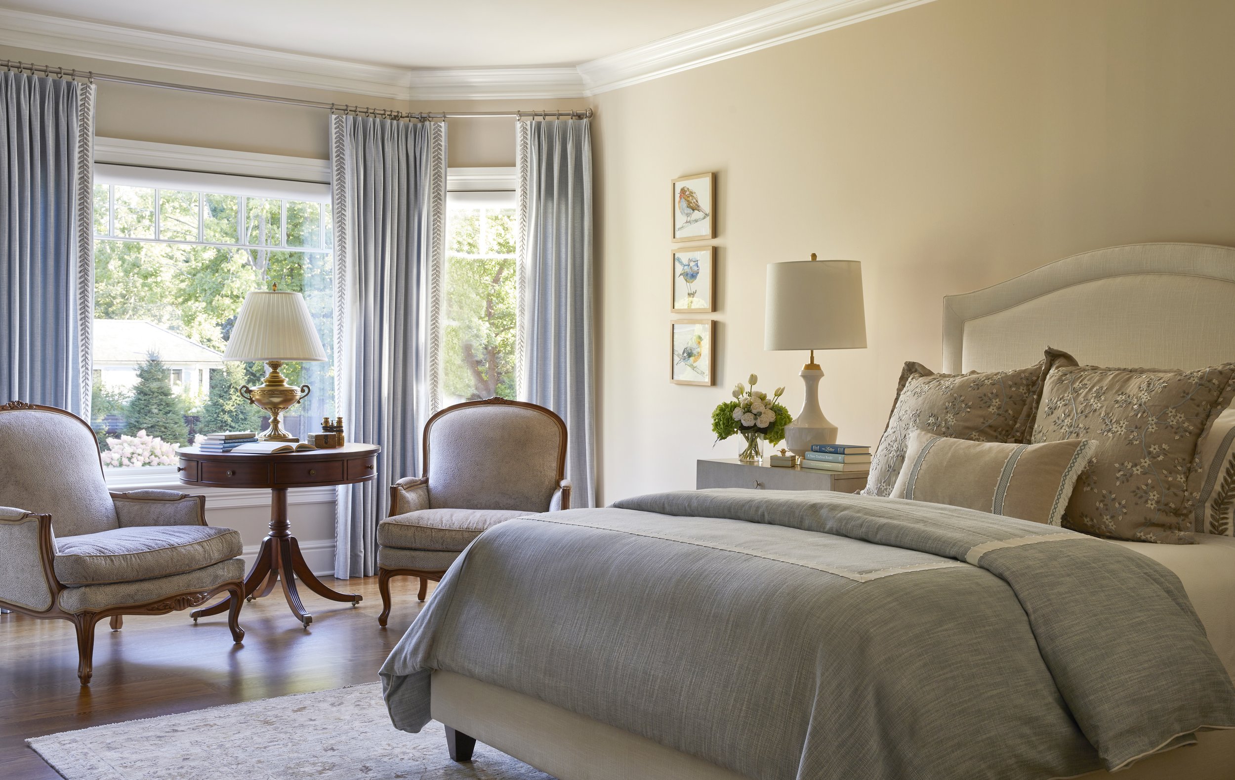Cozy bedroom with a large bed, two wooden armchairs, a round table with a lamp, and windows with blue curtains letting in natural light.