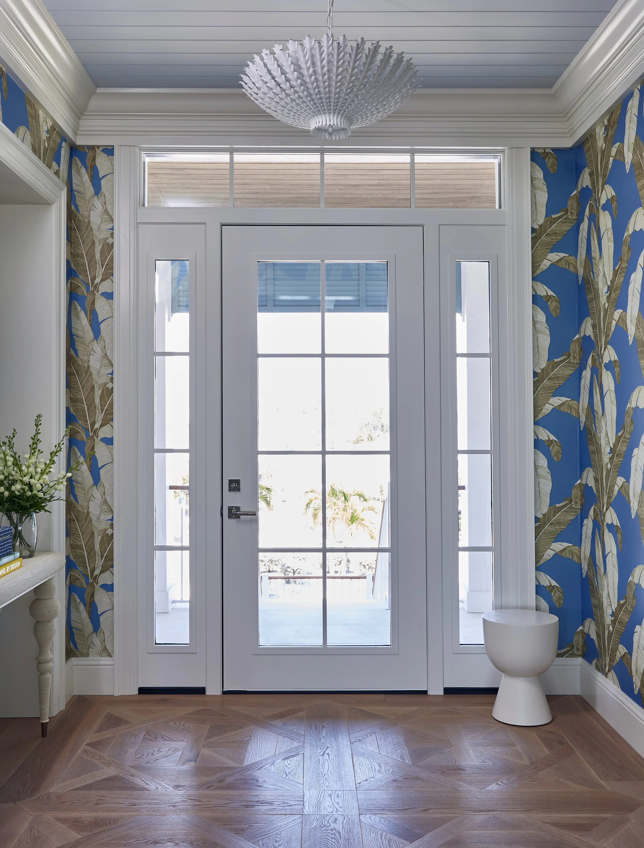 Front door with glass panels, flanked by two narrow windows, in a room with tropical patterned wallpaper, a chandelier, wooden flooring, a side table with flowers, and a small white stool.