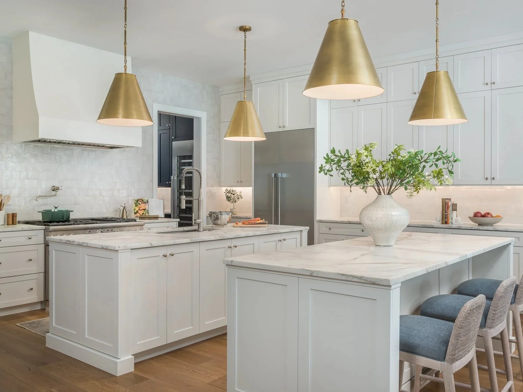 Modern white kitchen with gold pendant lights, marble countertops, a large central island, and a vase with green leaves.