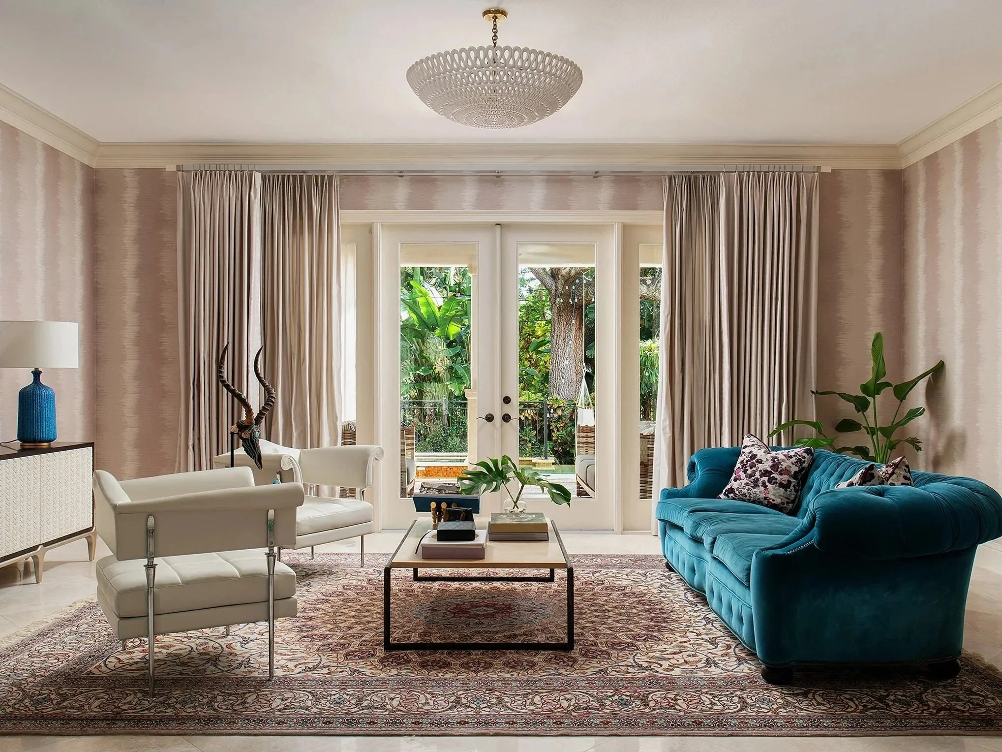 Living room with beige wallpaper, white curtains, beige ceiling, and a large window with glass doors looking out onto greenery. Furniture includes a white lounge chair with a matching ottoman, a turquoise velvet sofa with patterned throw pillows, a wooden coffee table with books and a leaf-shaped decor, a white sideboard with a blue vase and lamp, and potted plants. A large, intricate rug covers the floor, and a round chandelier hangs from the ceiling.