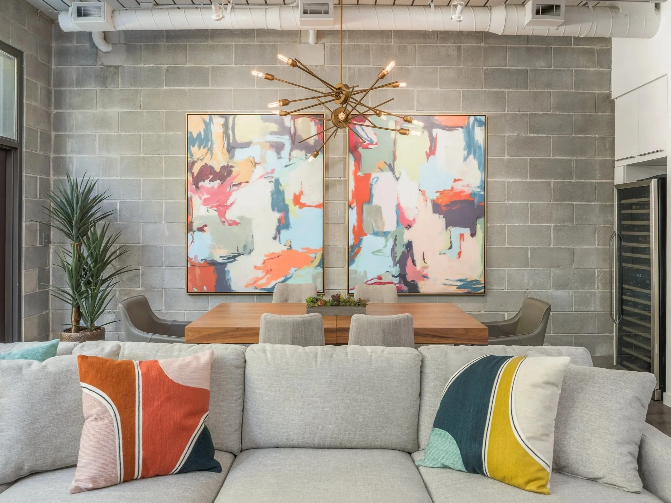 Living room with gray couch and colorful pillows, dining area with two modern chairs, green plant, abstract artwork, and a geometric chandelier on a gray cinder block wall.