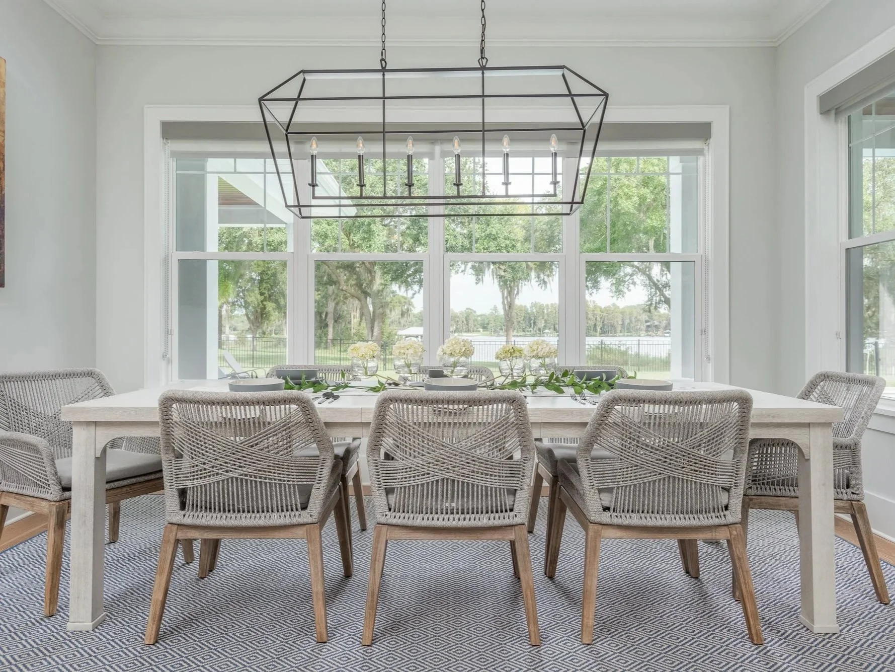 A bright dining room with a large white table, eight gray woven chairs, and a modern rectangular chandelier. Large windows reveal a view of trees and a body of water outside.