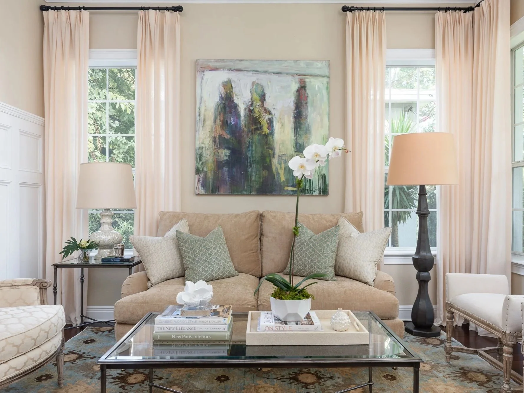 Living room with beige sofa, glass coffee table with magazines, potted orchid, and decorative objects. Cream curtains flanking windows, colorful abstract painting on beige wall, lamps on side tables, and armchair with white upholstery.