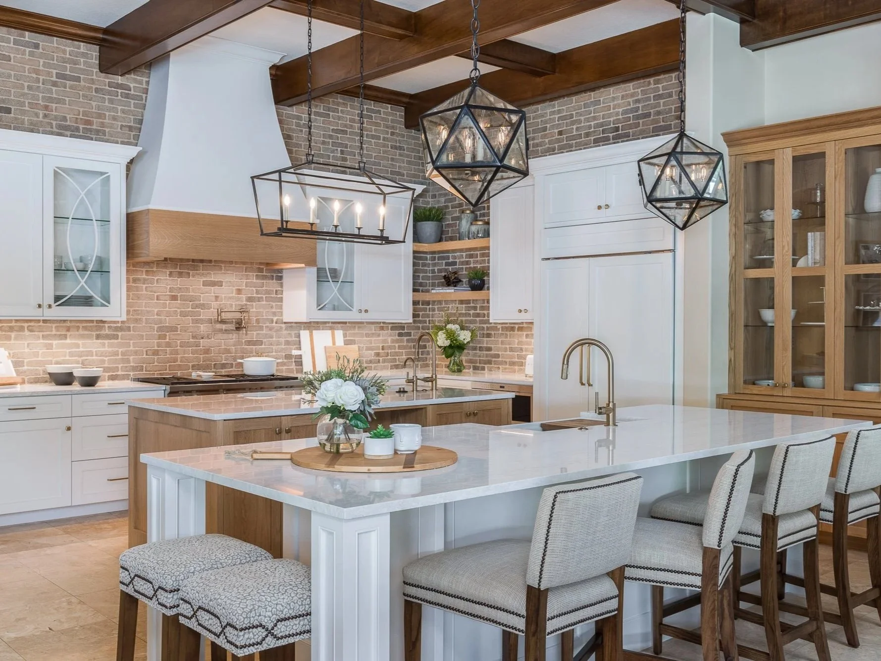 Modern kitchen with white cabinets, a large white island with seating, a brick wall backsplash, and wooden accents. Hanging geometric light fixtures above the island.