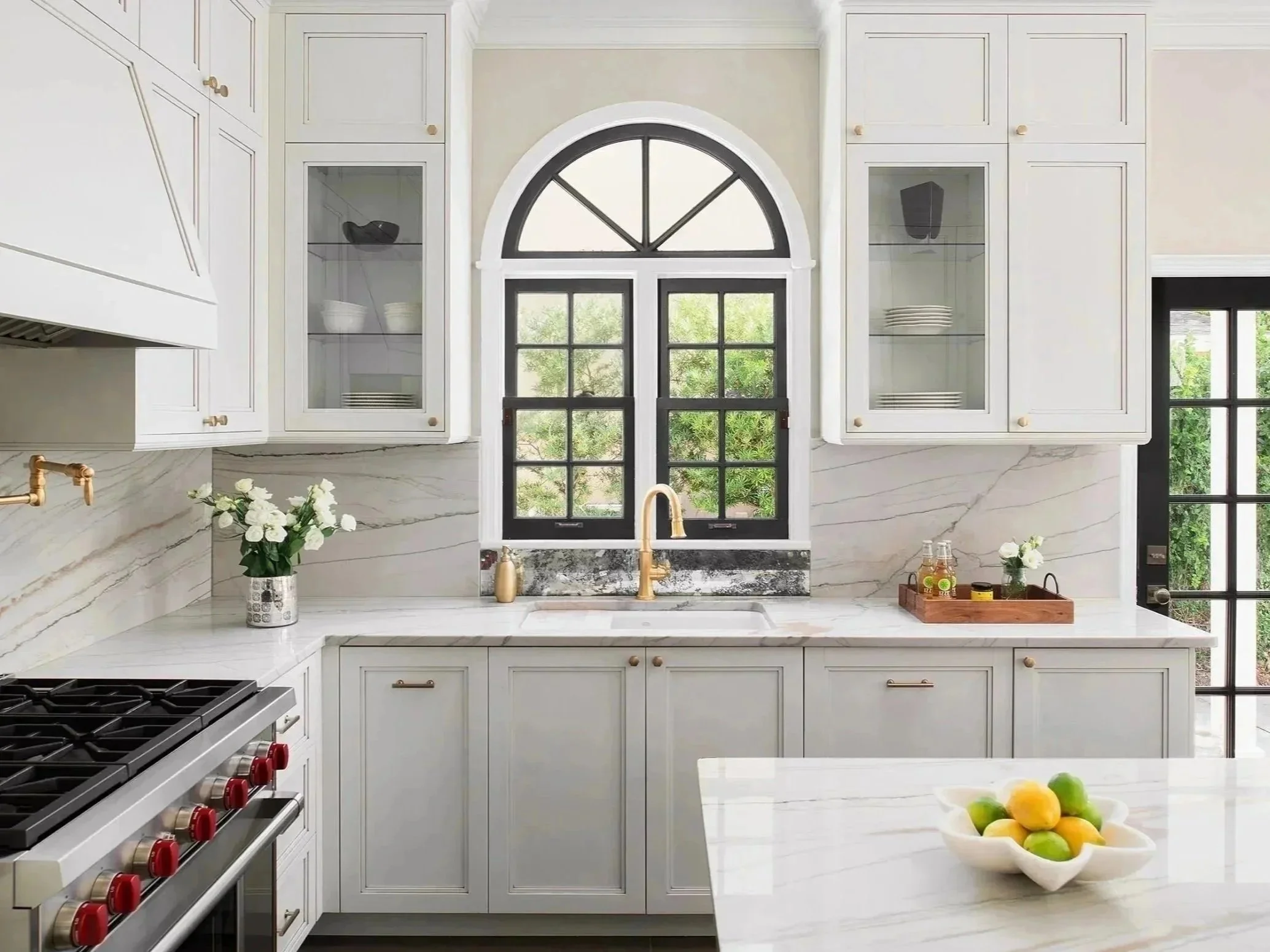 Bright white kitchen with marble countertops, cabinets, and backsplash. A large window above the sink with black framing, decorated with flowers and small decorative items. A bowl of lemons and limes on the island counter. Kitchen appliances and dishes are visible.